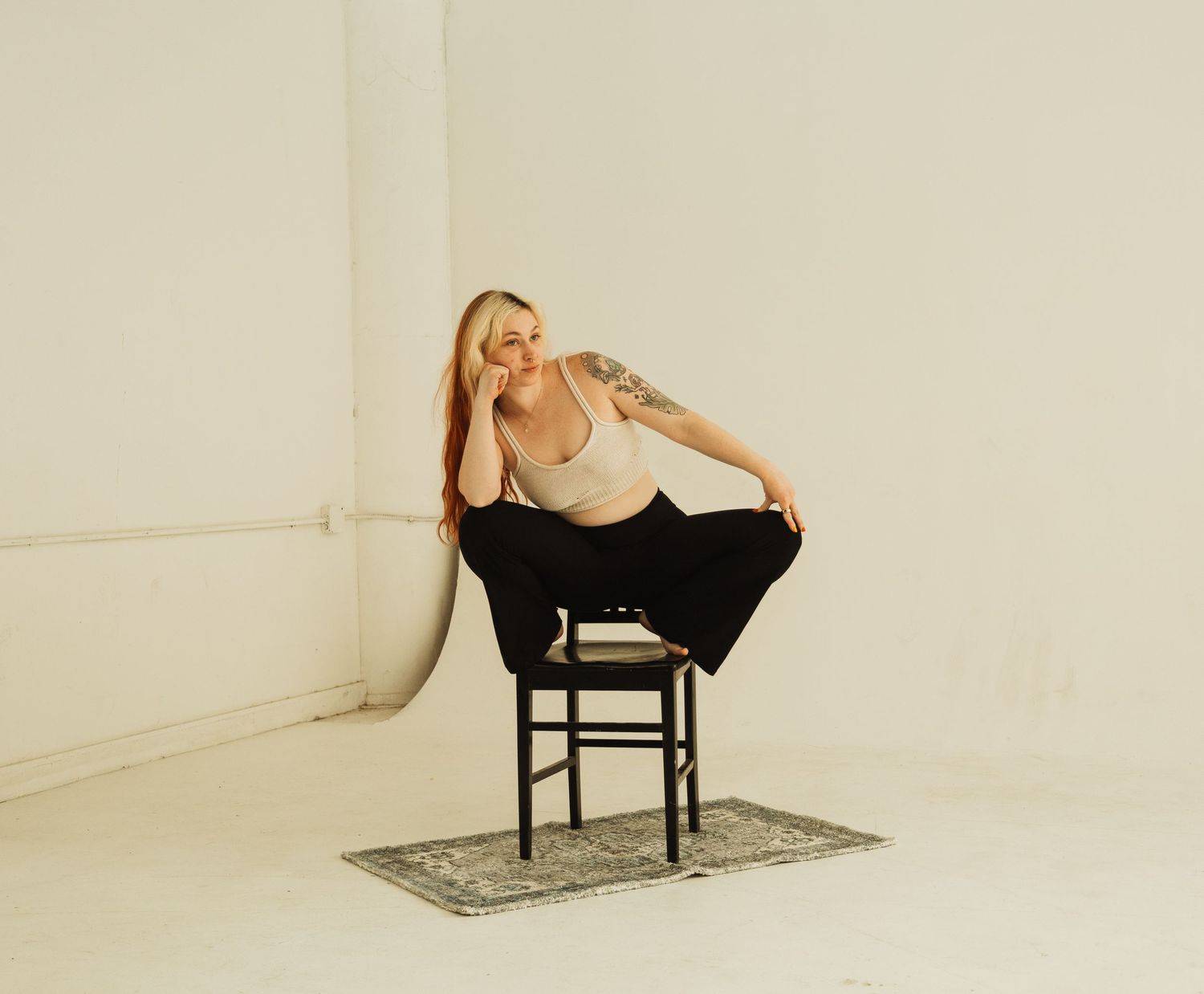 A model in casual attire strikes a dynamic pose while perched on a black chair in a minimalist studio setting.