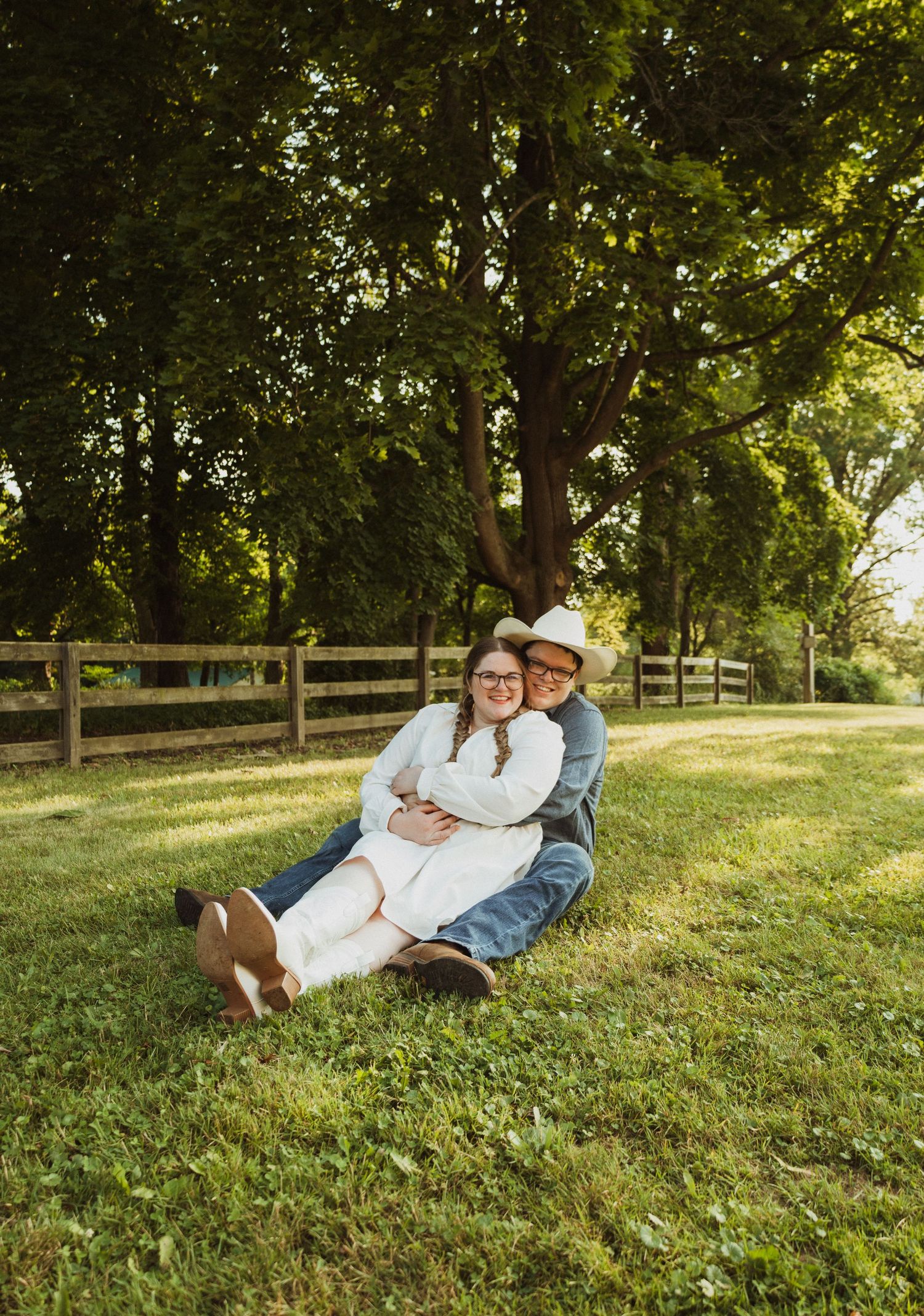 Intimate outdoor photoshoot on a grassy slope with wooden fence and trees in background.