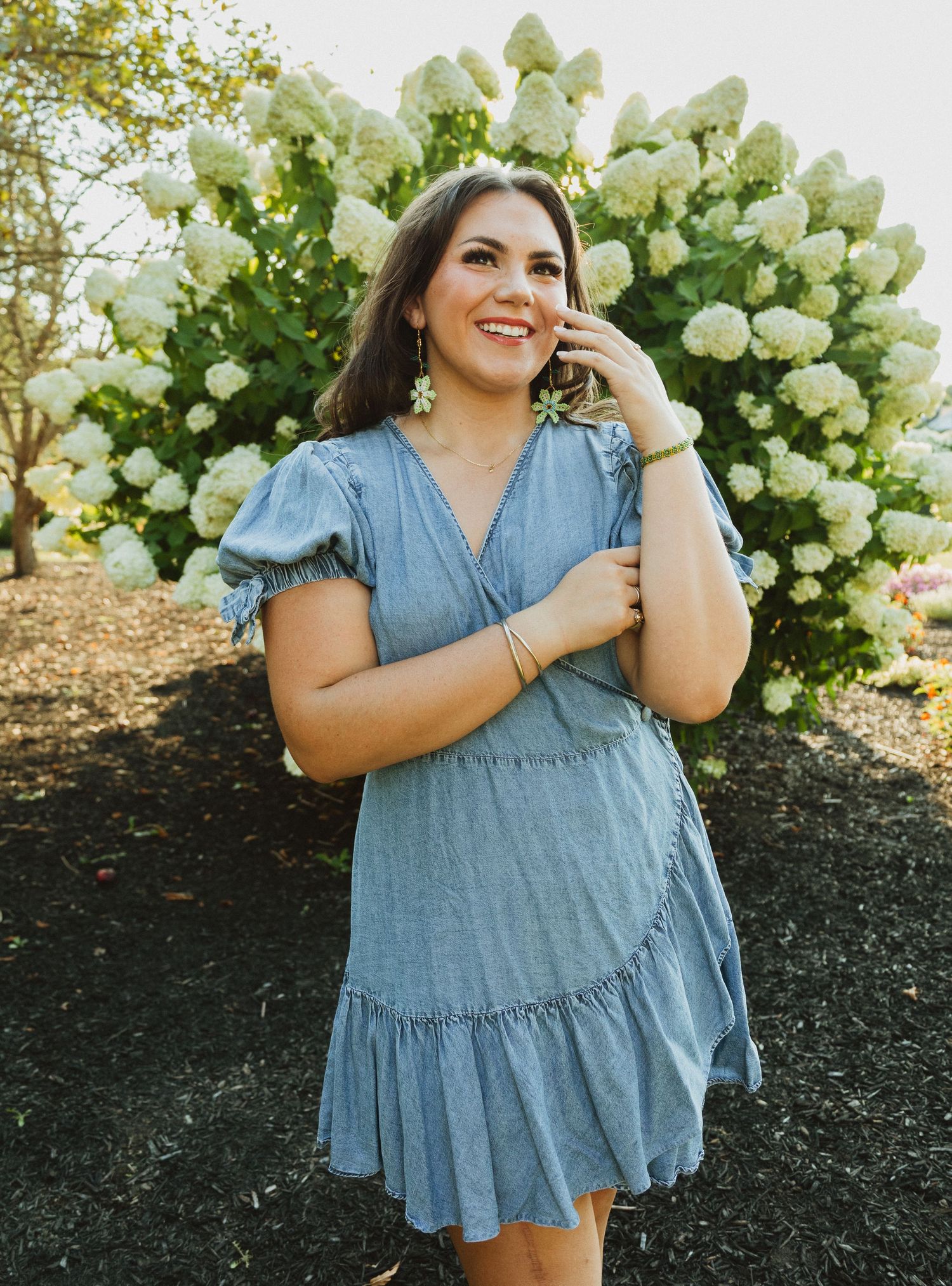 Woman in a blue denim ruffle dress poses against white flowering bushes in an outdoor garden setting.