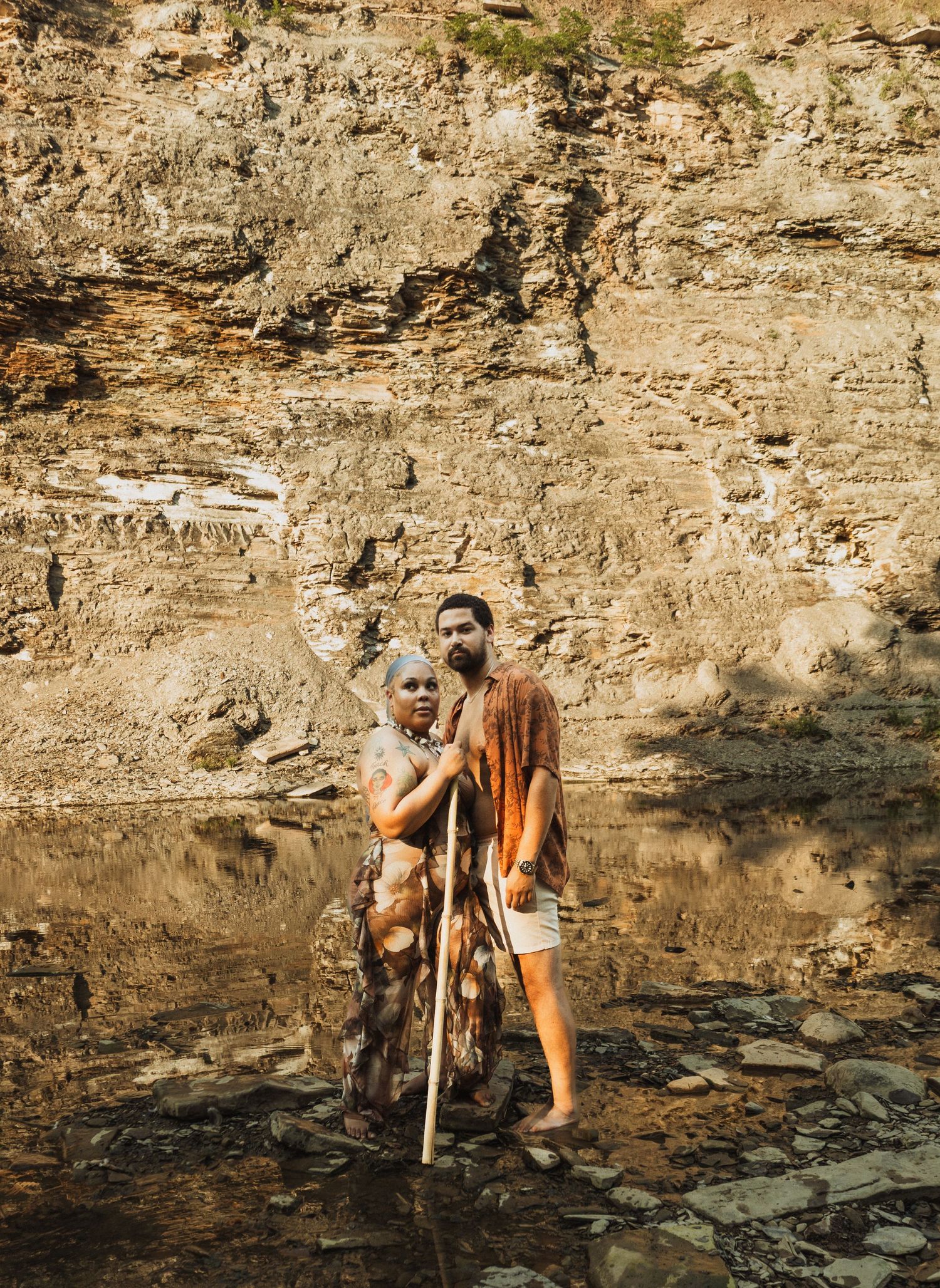 A scenic portrait by a rocky cliff face with a person holding a large dog while standing on stones near a shoreline.