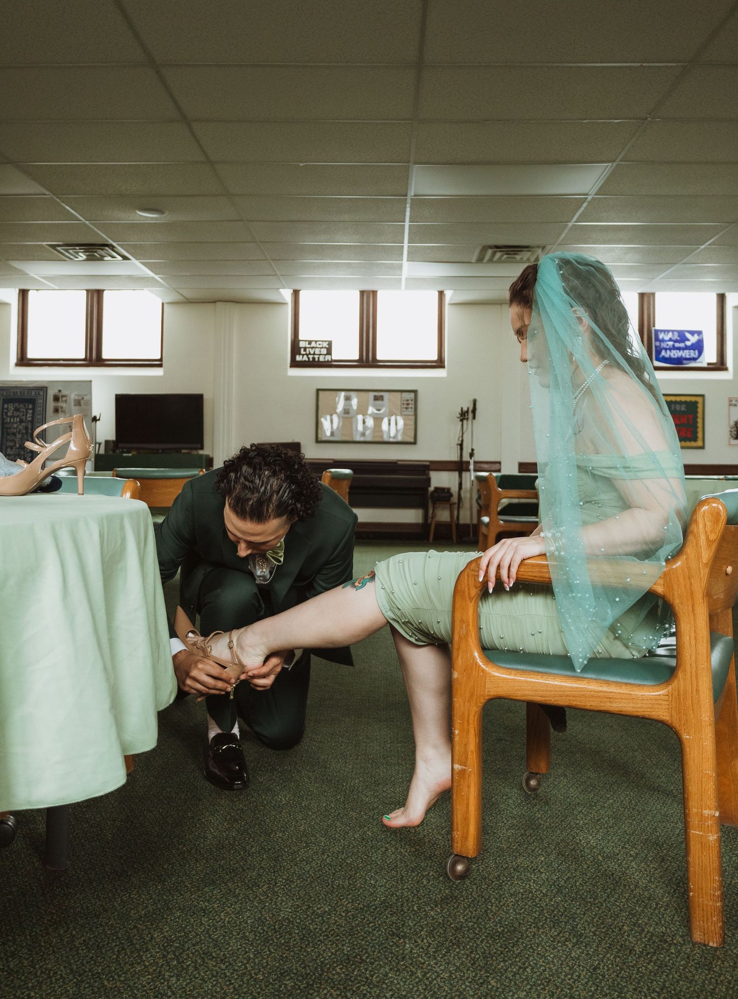 Groom kneeling to put shoes on barefoot bride in green dress and teal veil before ceremony.