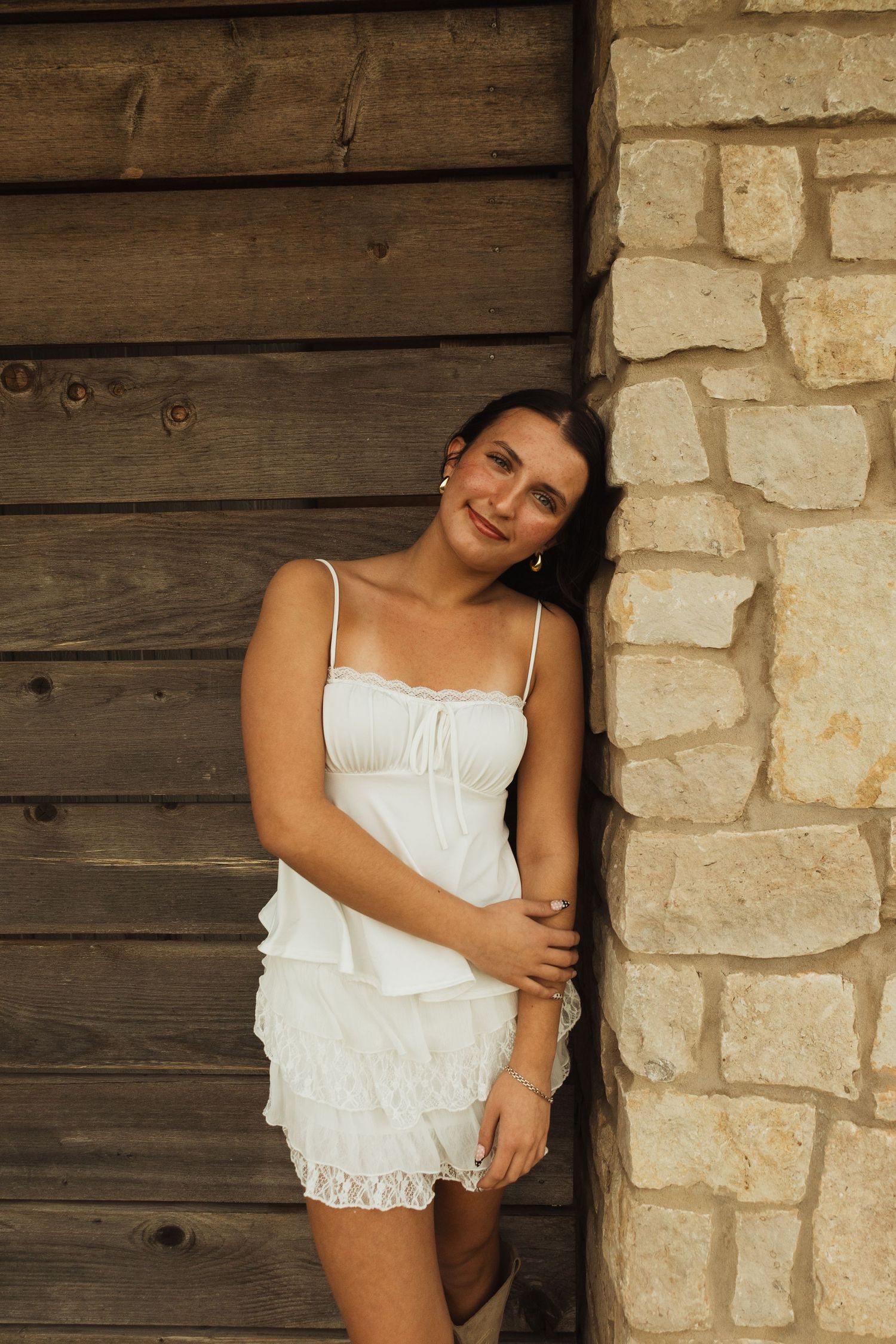Young woman in white lace romper leaning against stone wall and wooden barn backdrop, smiling at camera.
