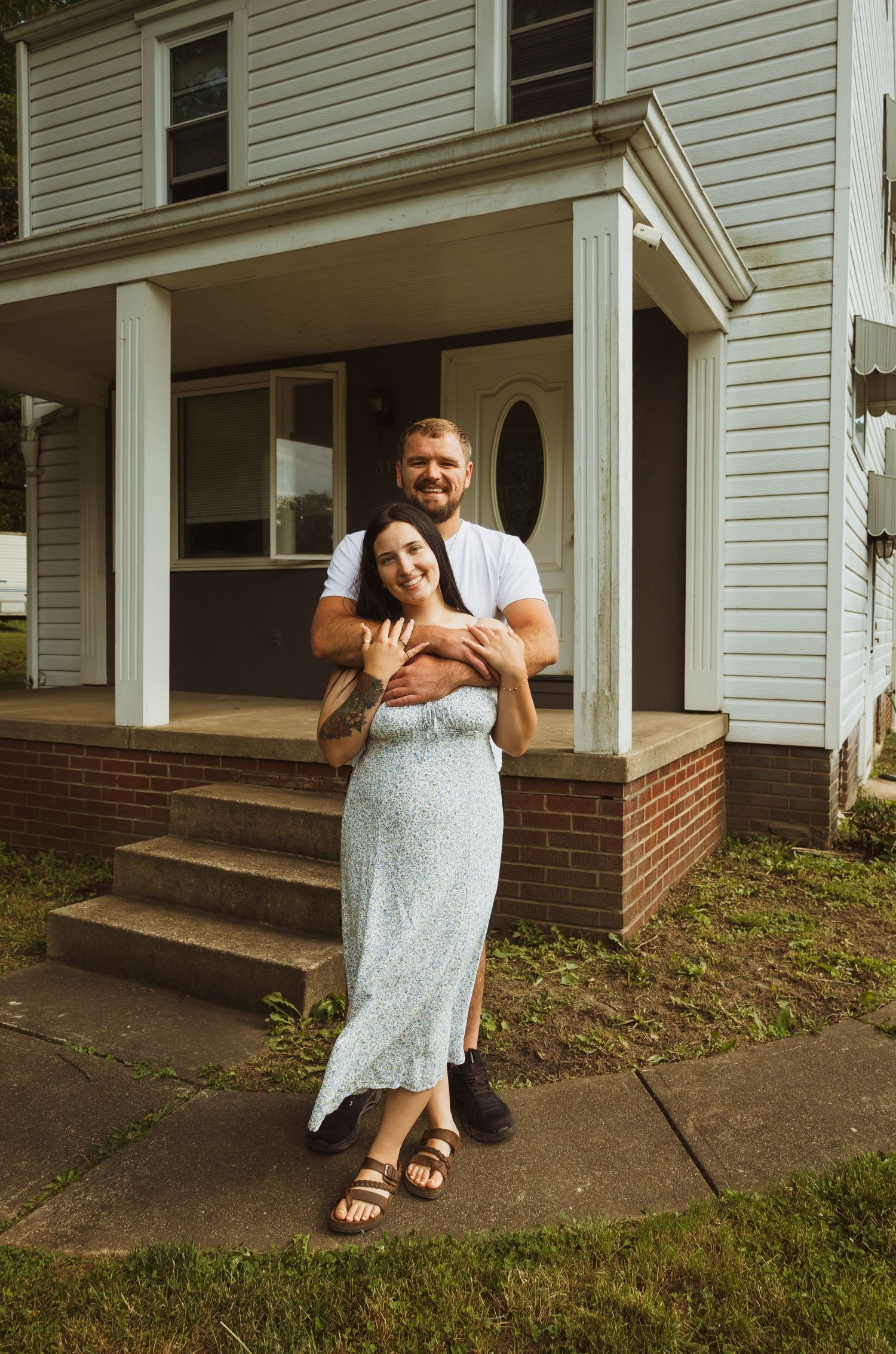A smiling couple embraces in front of a white house porch, woman in floral dress, man in white tee, standing on a sidewalk.