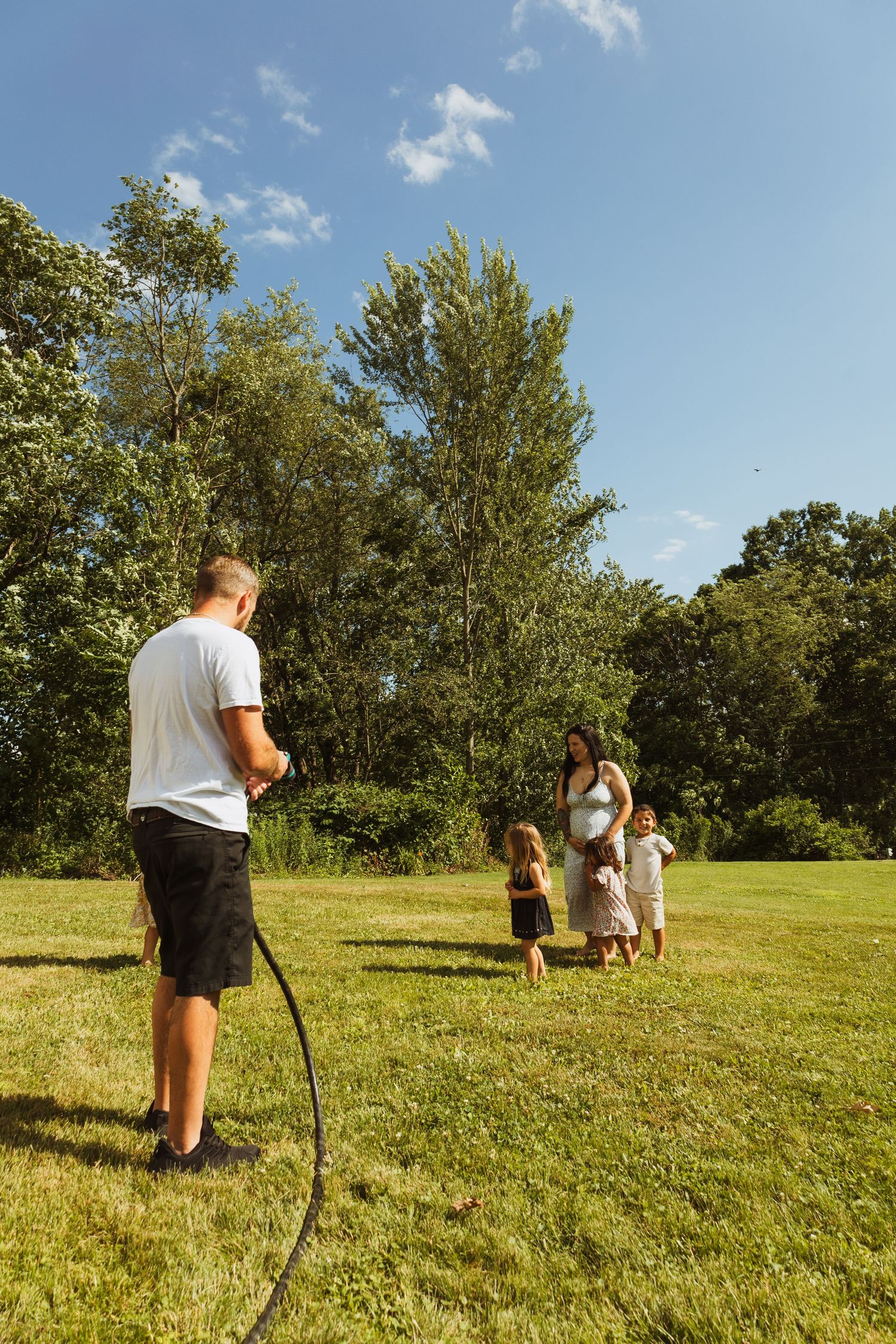 Family enjoying a sunny day outdoors, with a man holding a garden hose while children and a woman stand on a green lawn.