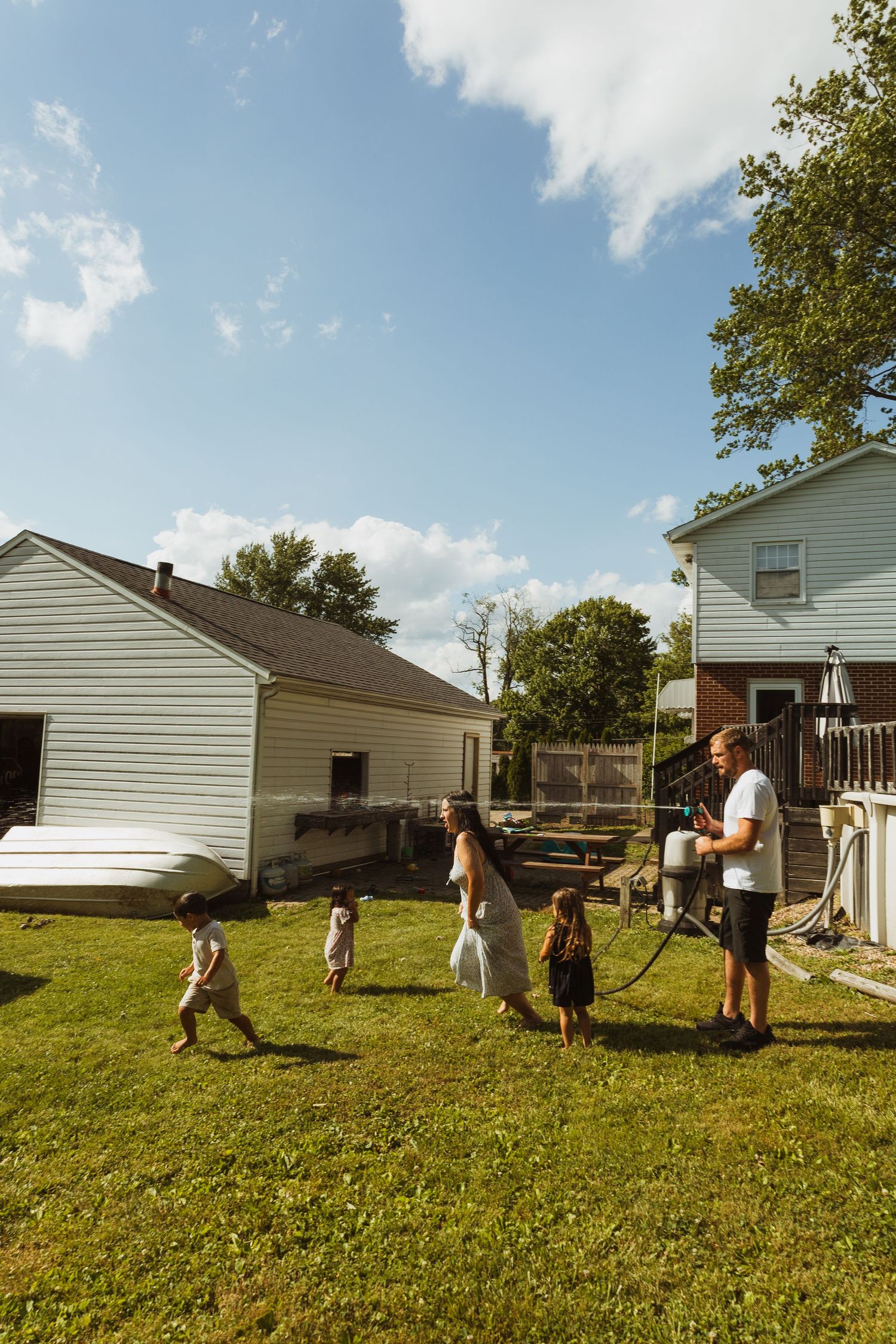 Family playing in backyard on sunny day, with children running on green grass near white suburban homes.