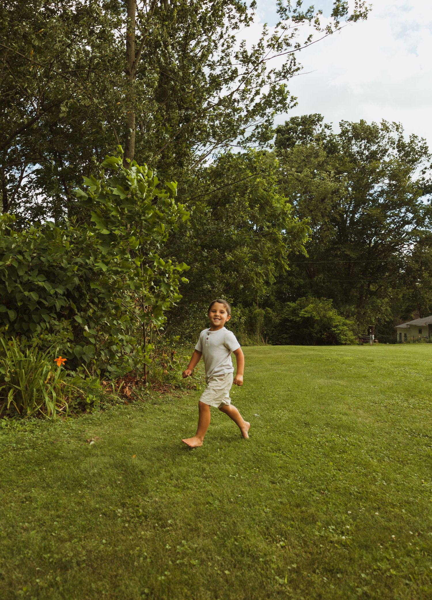 A barefoot child runs joyfully across a lush green lawn surrounded by trees on a sunny day.