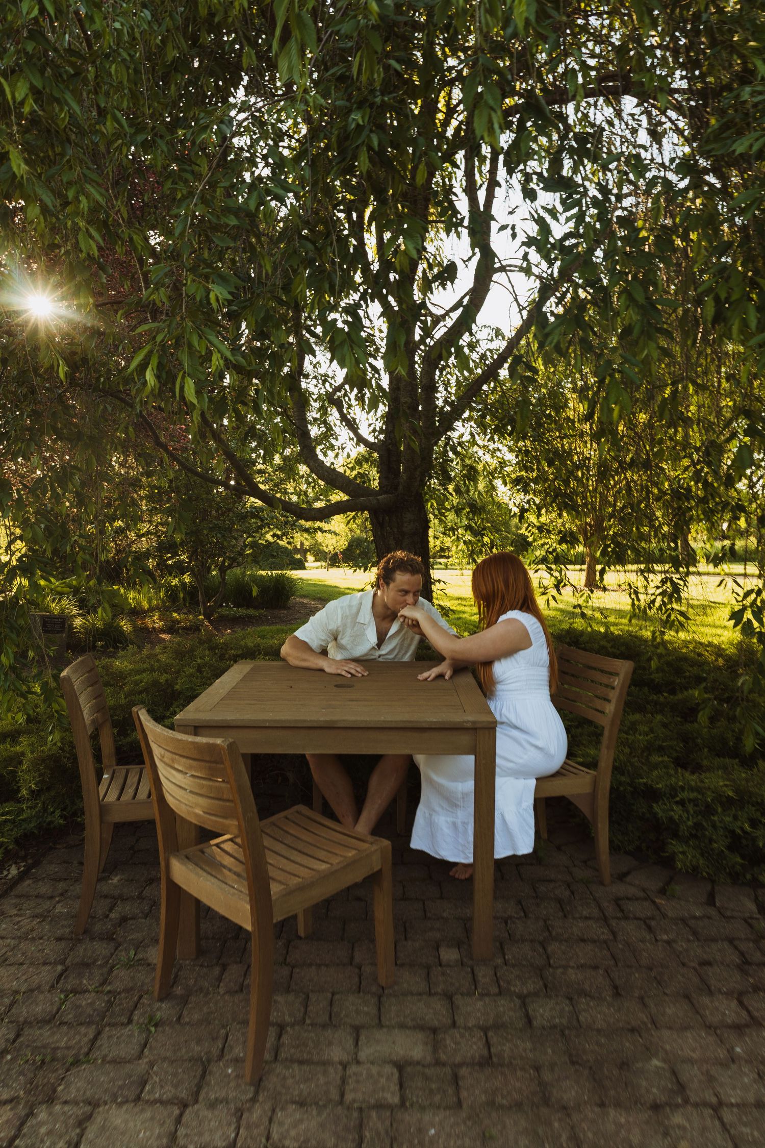 Couple sitting together at a wooden outdoor patio table surrounded by lush green trees in warm sunset light.