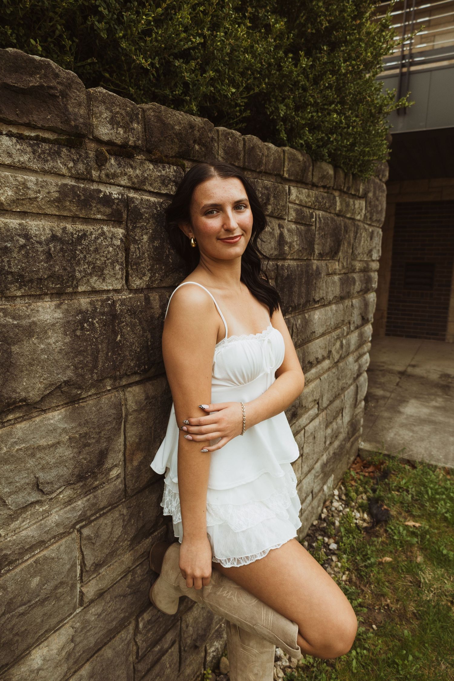 Young woman in white ruffled romper leaning against stone wall outdoors, smiling confidently.