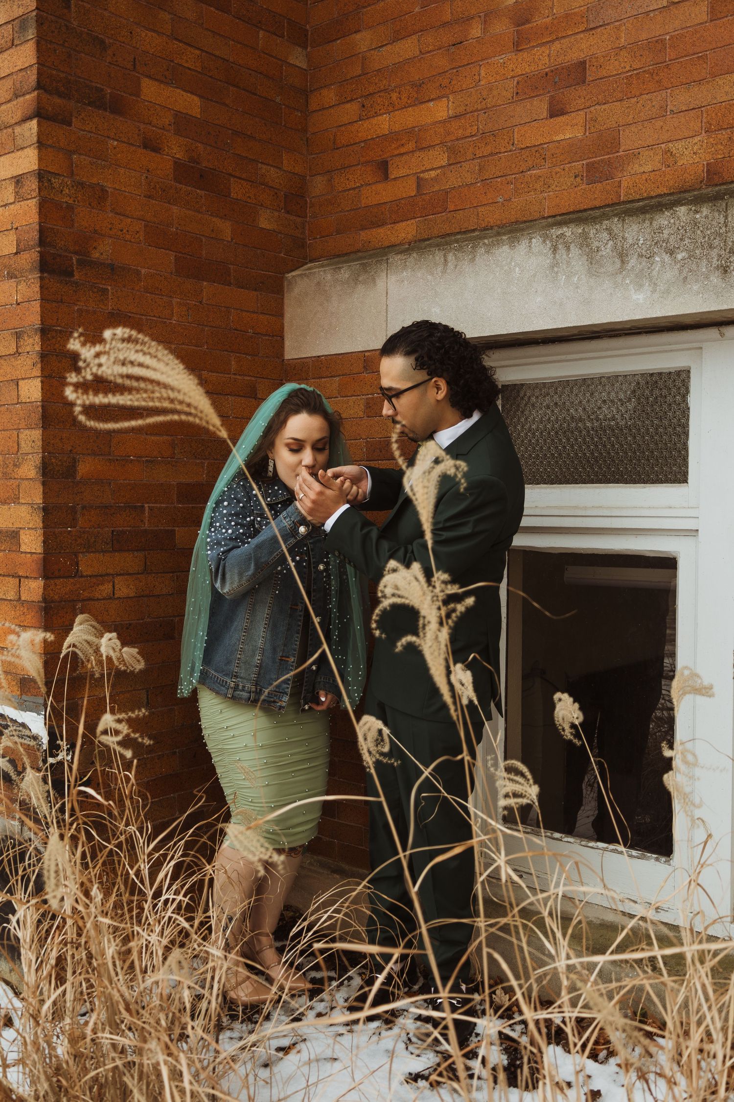 Couple sharing a tender moment outside a brick building surrounded by dried winter grasses and snow.
