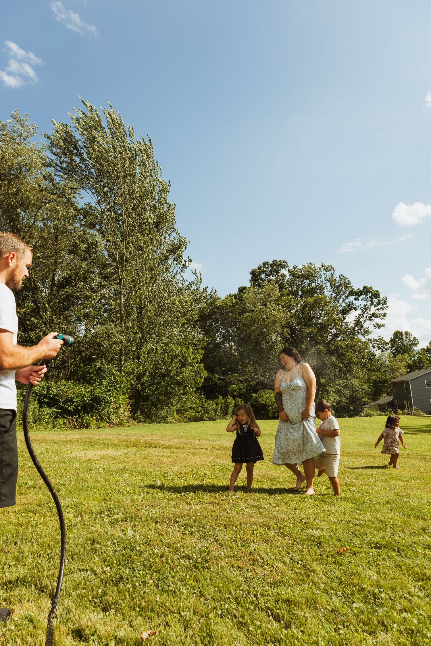 Family playing with garden hose in sunny backyard, man spraying water at woman and children running on green grass.