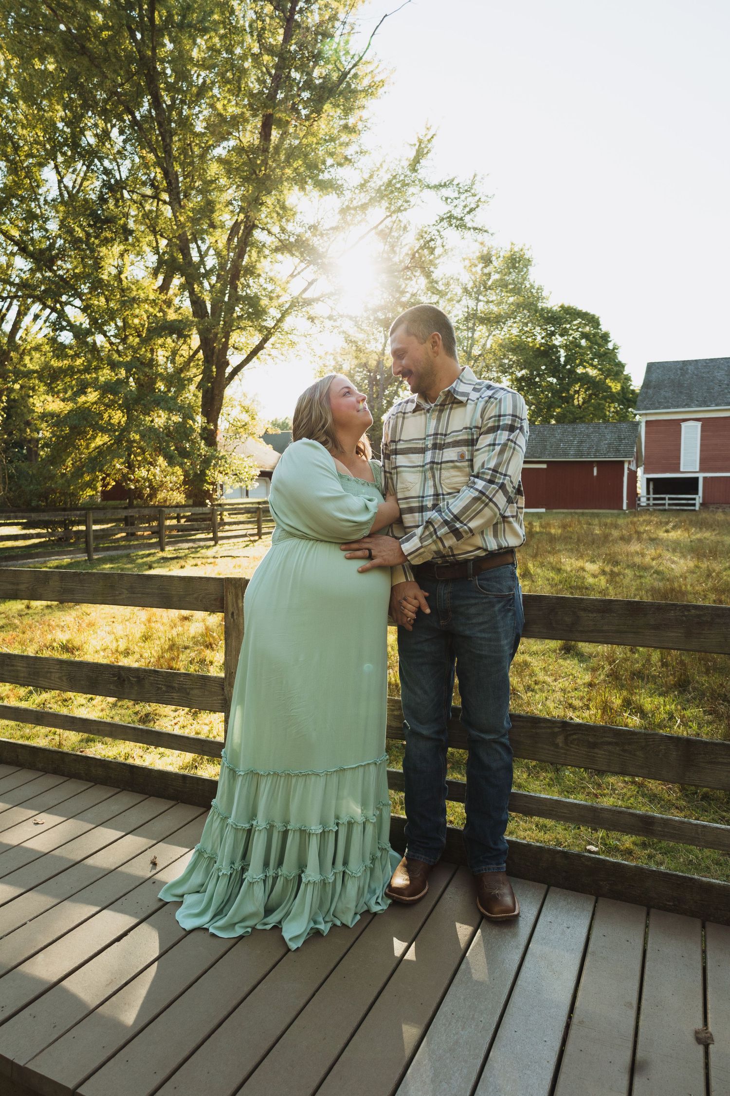 A couple embraces tenderly on a wooden deck at sunset with a rustic barn and trees in the background.