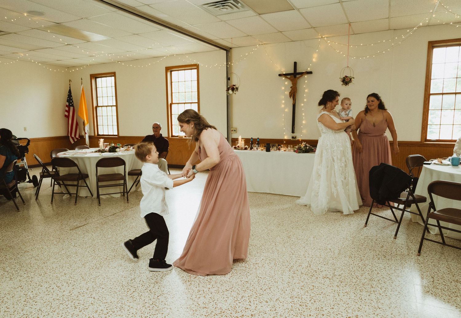 Wedding party dancing and celebrating in a spacious church reception hall with tiled floors.