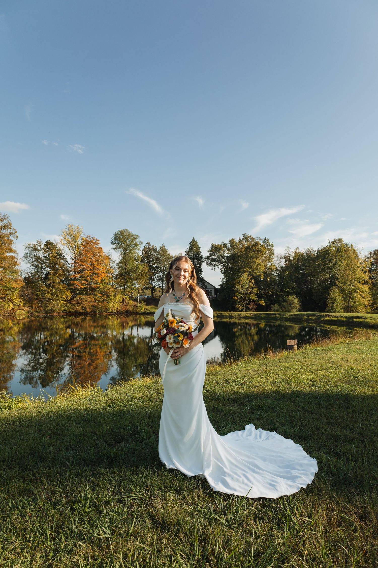 White wedding dress flowing in the breeze against a scenic lakeside backdrop with autumn trees.