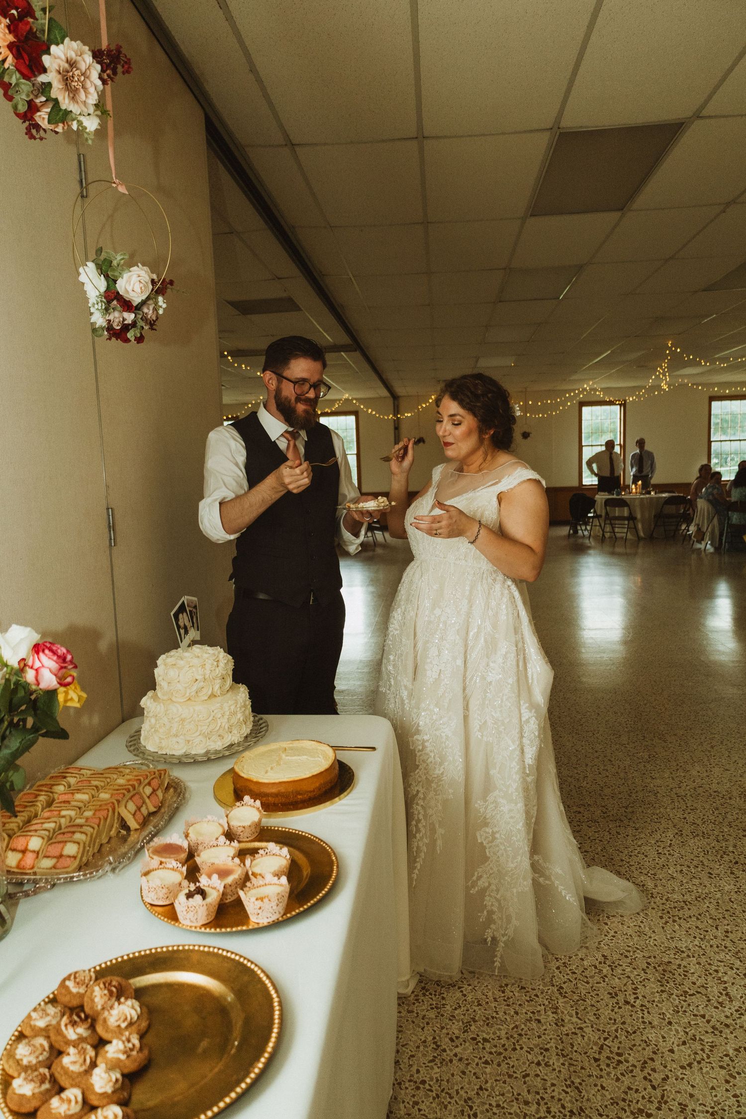 A couple shares a moment while tasting cake at their wedding reception dessert table with various sweets displayed.