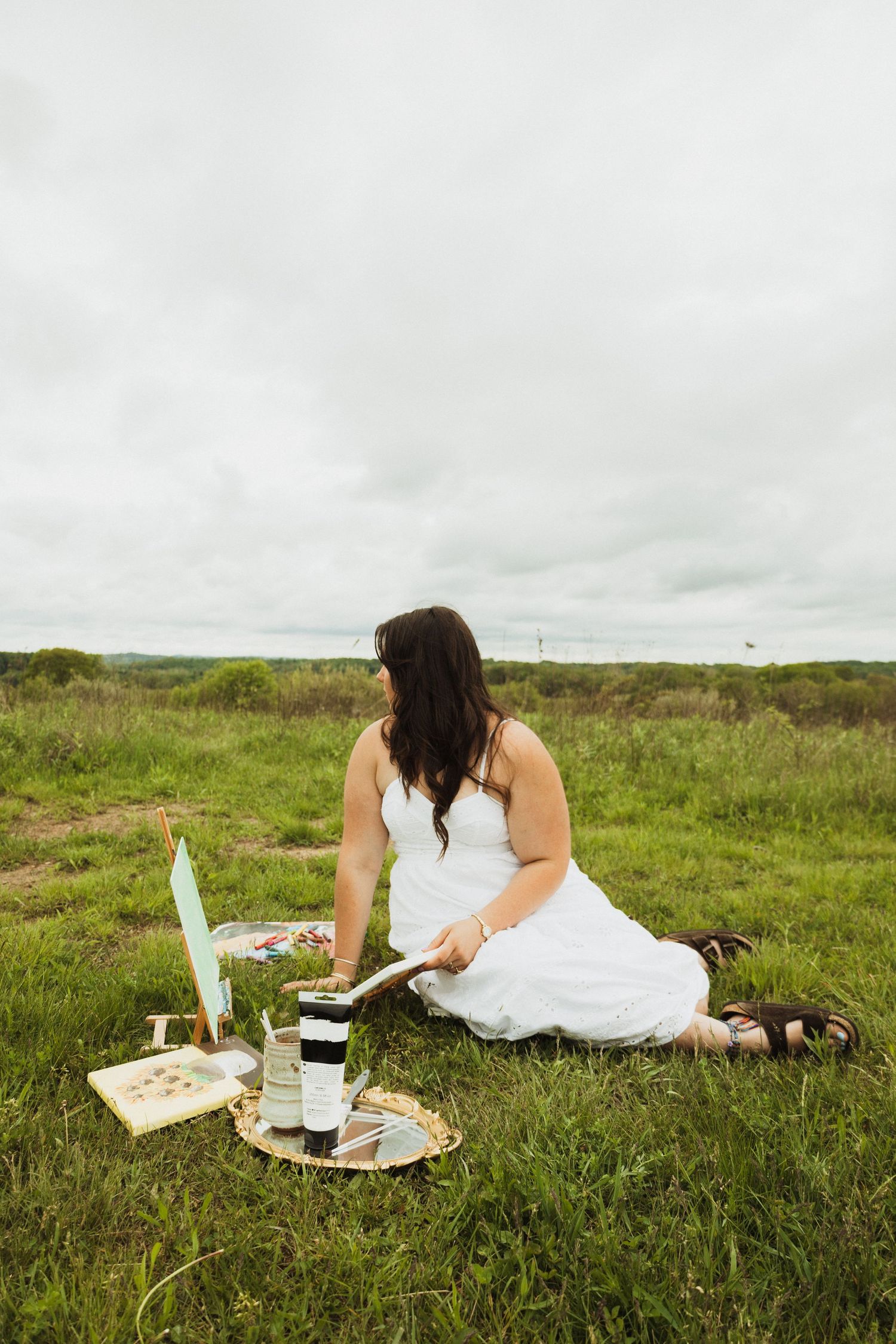 Woman in white dress sits on grass for picnic with wine, candle and journal on cloudy day.