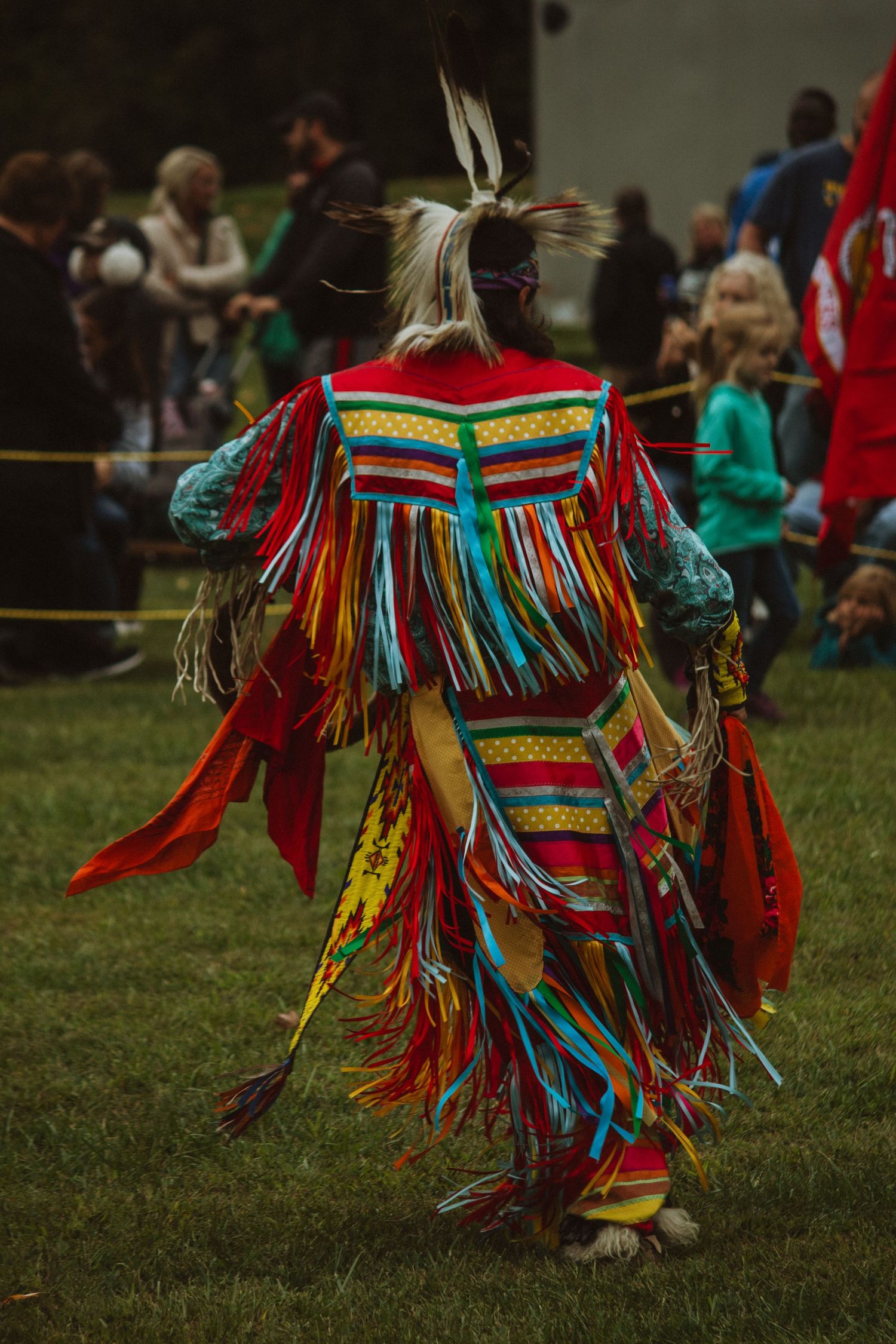 A vibrant powwow dancer in colorful traditional regalia with feathers and ribbons performs at an outdoor gathering.