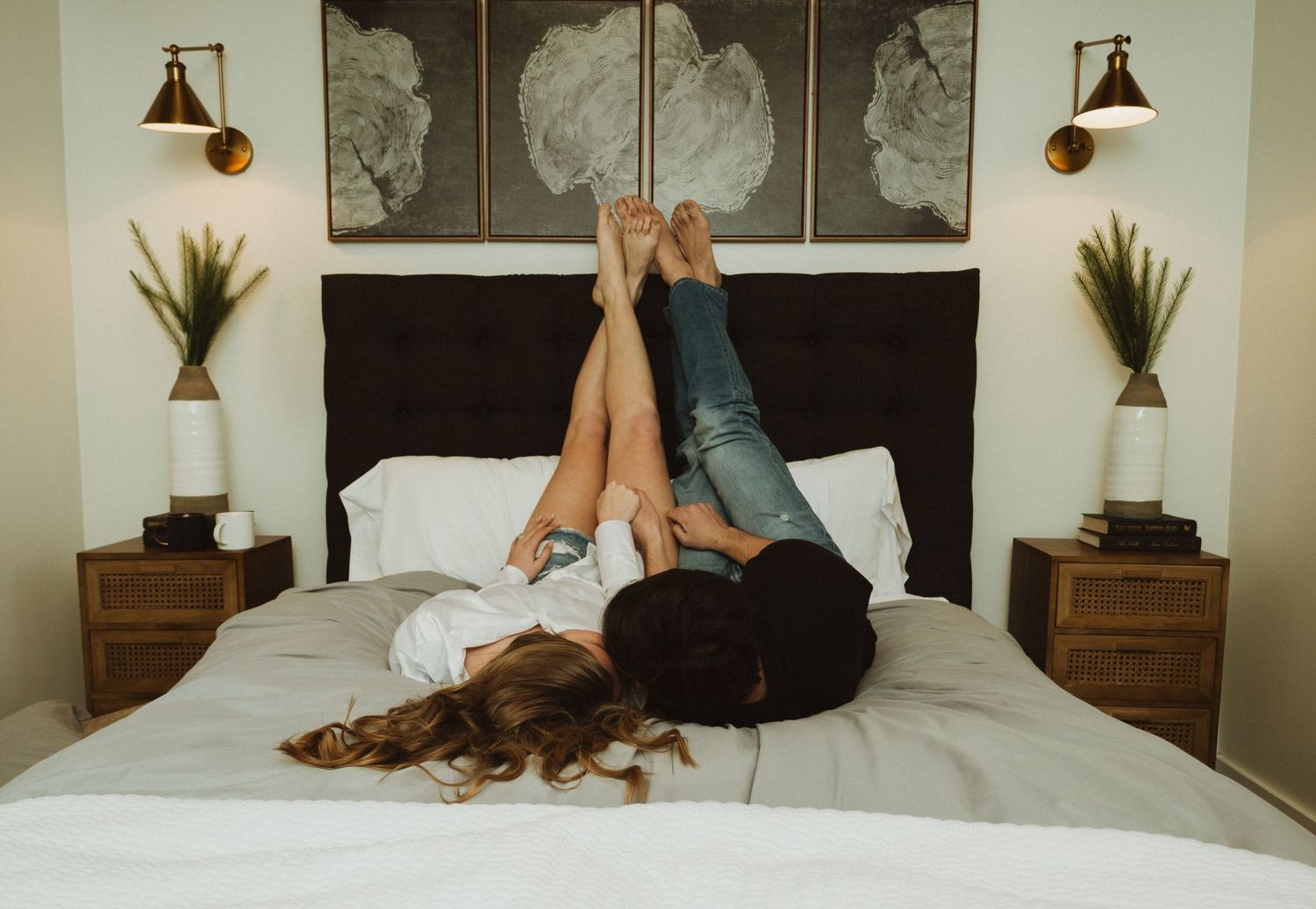 Two individuals lying on a bed with legs raised against a wall featuring cloud artwork and wooden bedside tables.