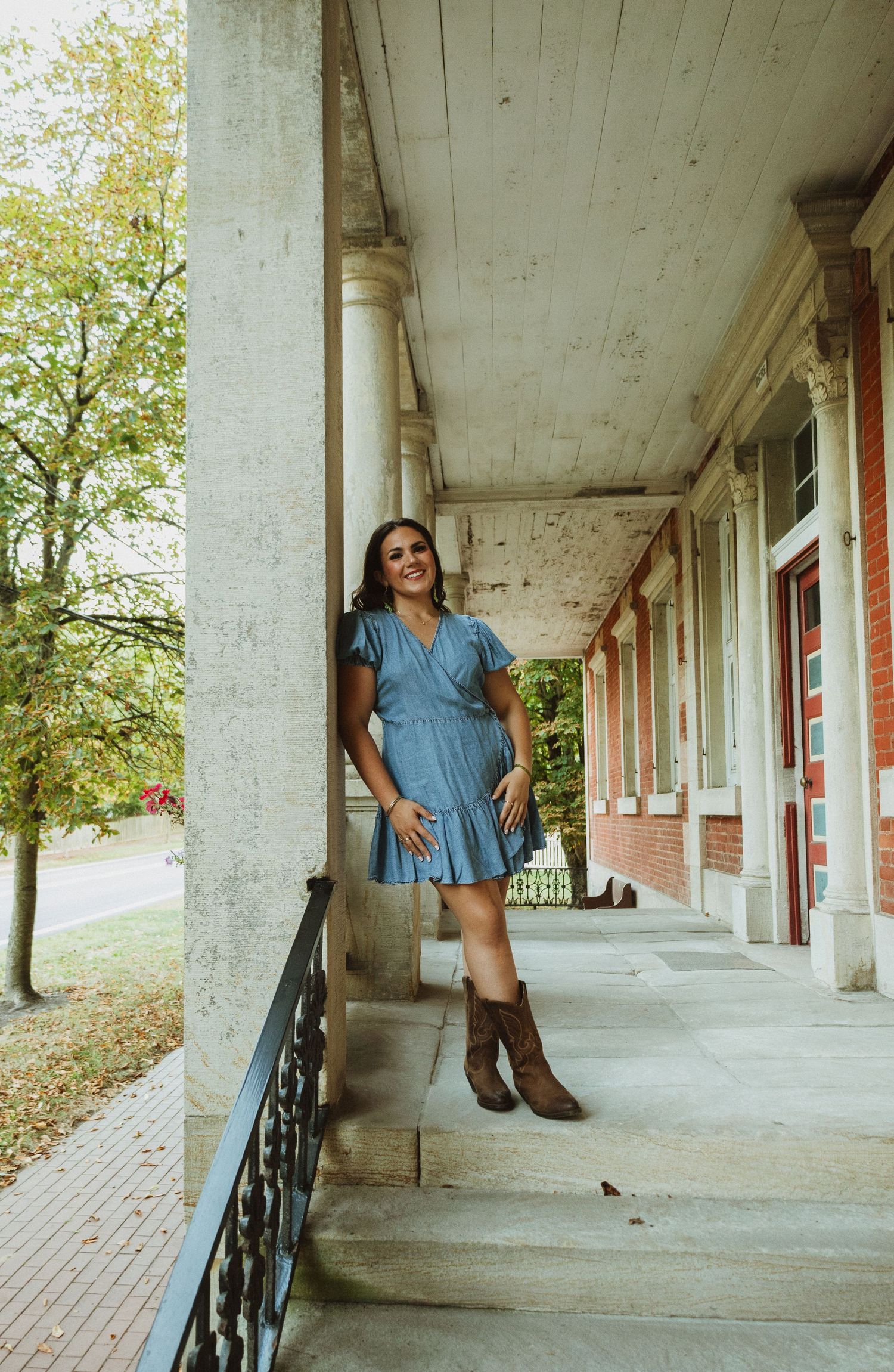 A person in a blue dress poses along a covered walkway with white columns and railings on a sunny day.