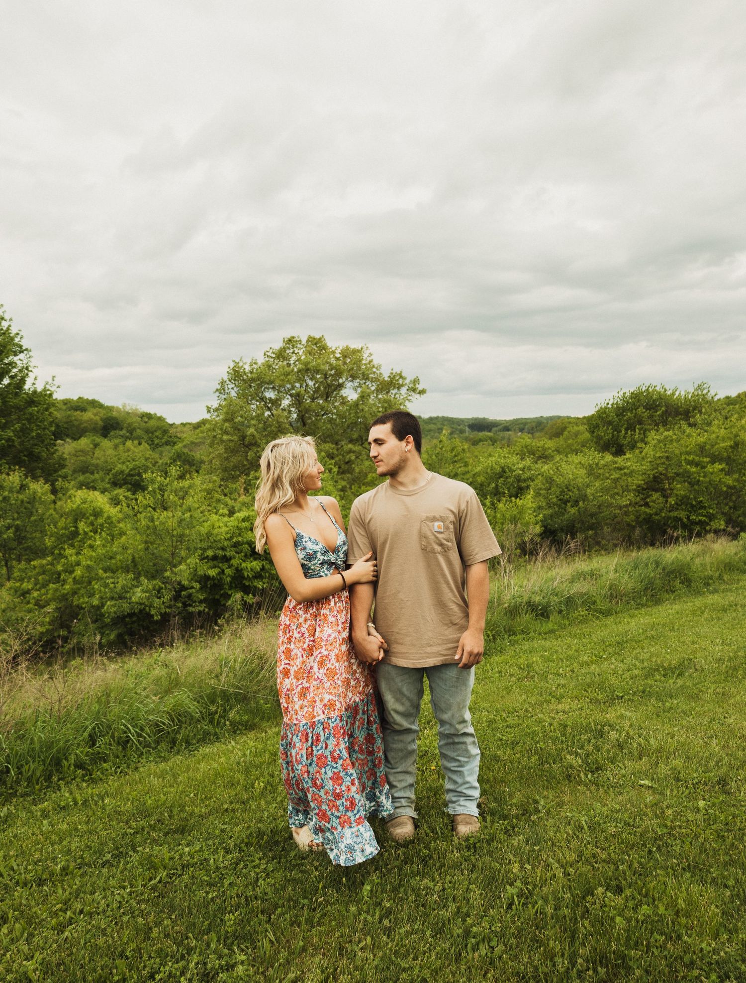 A couple stands together in a grassy field on an overcast day with lush greenery in the background.