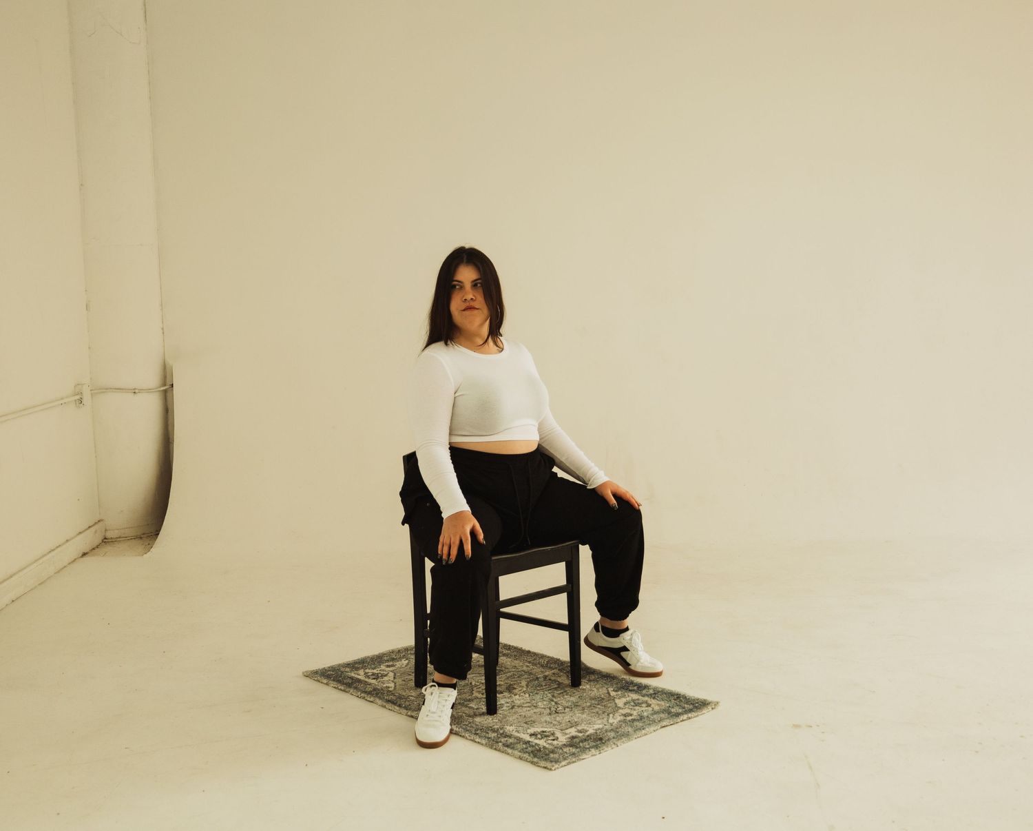 Someone wearing a white crop top and black pants sits on a wooden chair against a beige wall backdrop with ornate rug below.