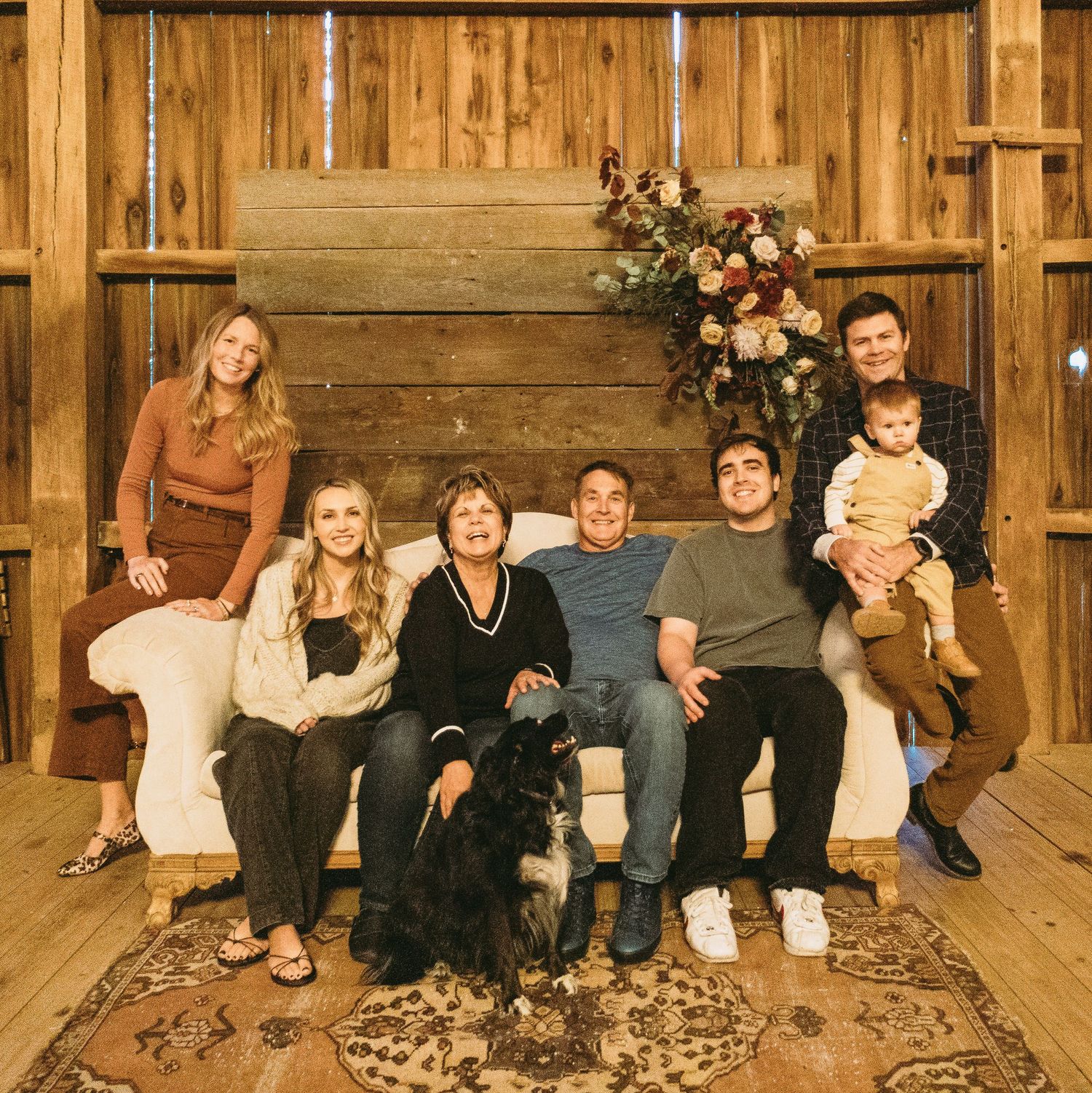 Indoor family gathering with pet dog on white couch against rustic wooden wall with floral decor.