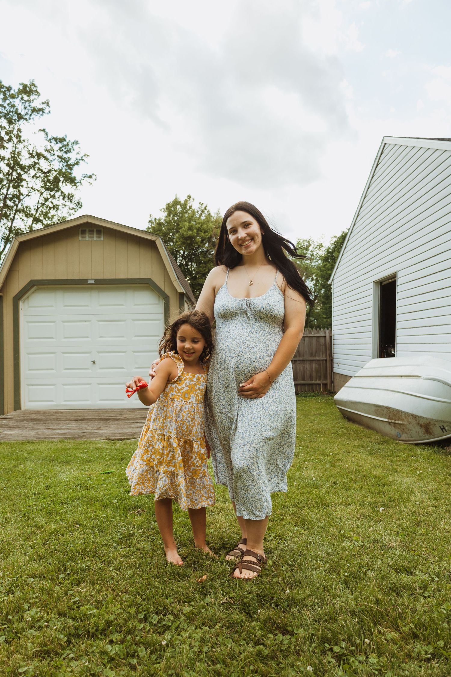 Pregnant woman in floral dress stands with young girl in yellow dress in backyard near garage on cloudy day.