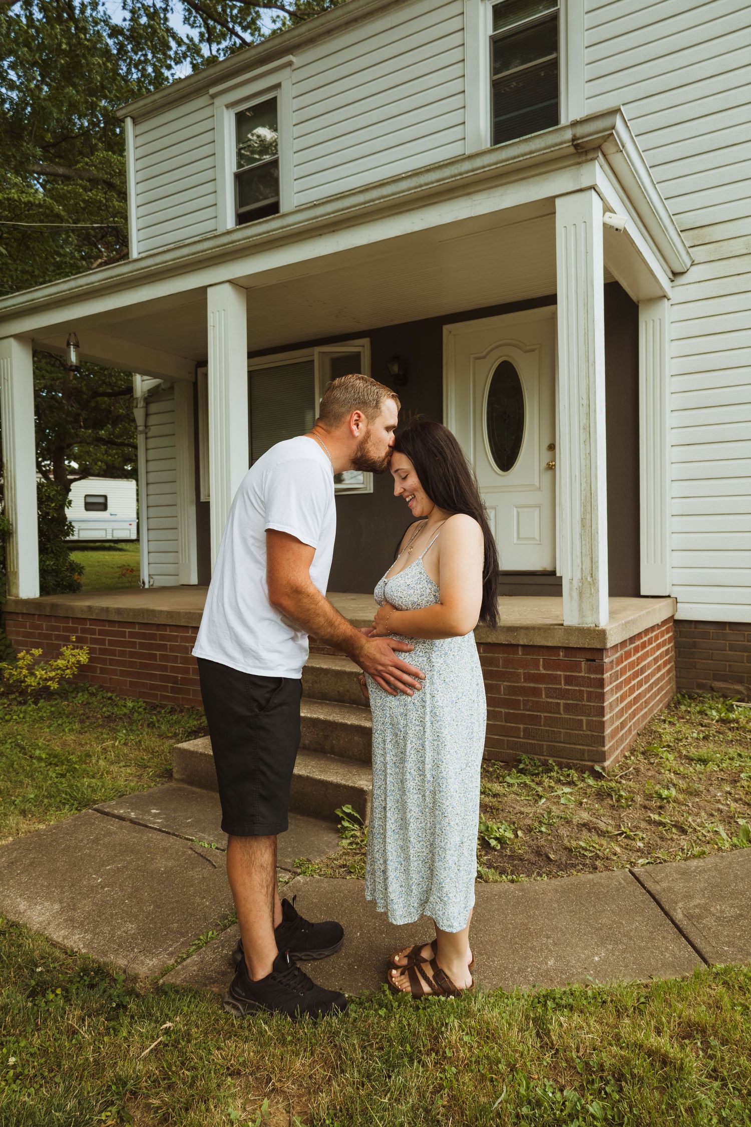 Couple holding hands in front of a white house porch, woman in floral dress smiling as man leans in close.