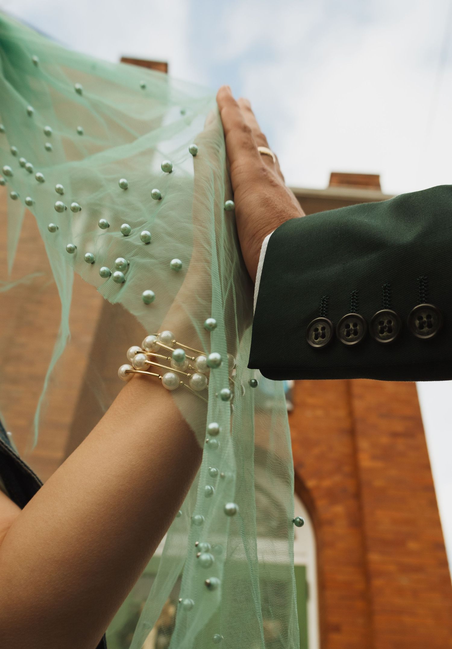 Close-up of bride's pearl bracelet and groom's dark suit sleeve holding teal pearl-adorned wedding veil outdoors.