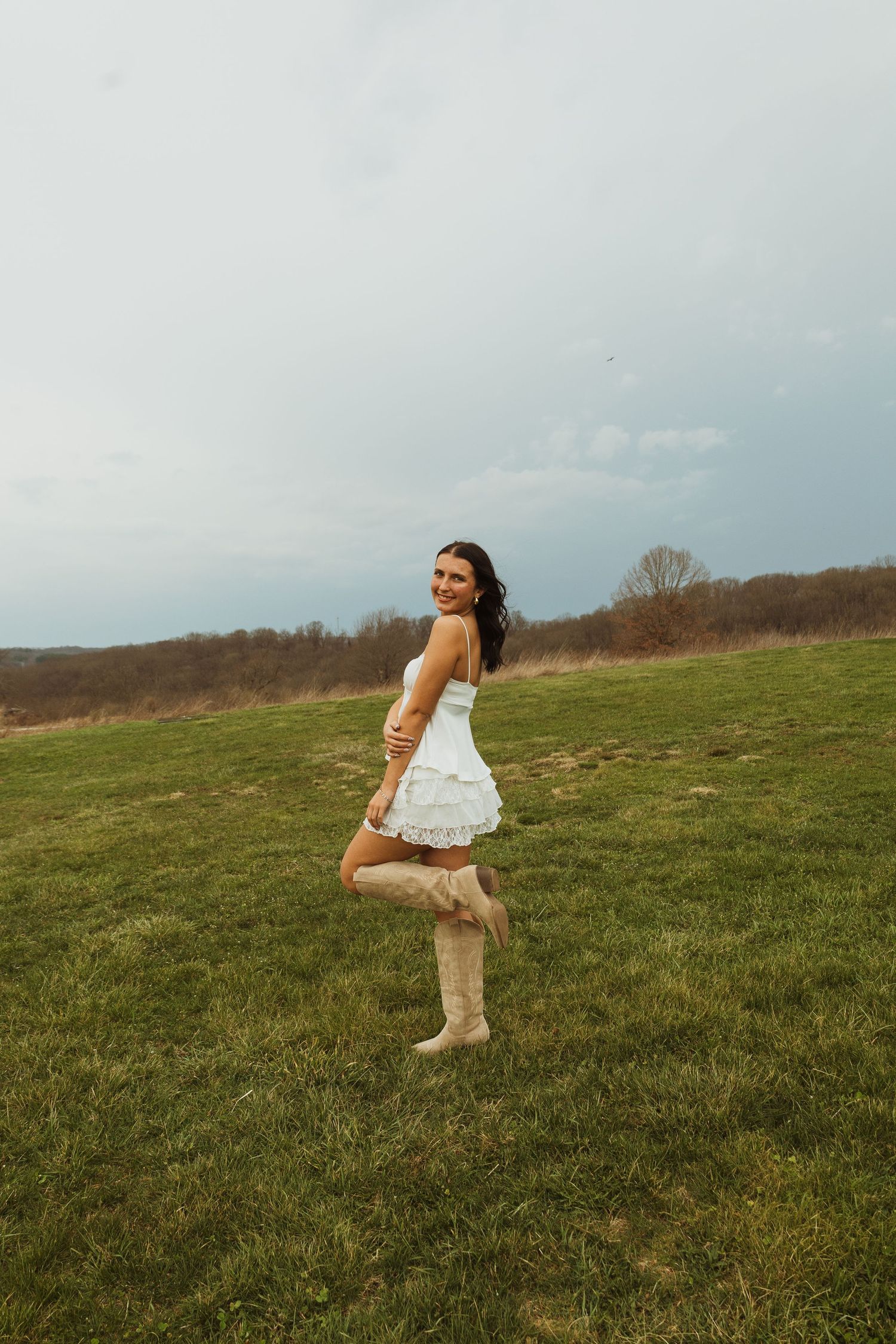 Young woman in white ruffled mini dress and cowboy boots posing on green field under cloudy sky.