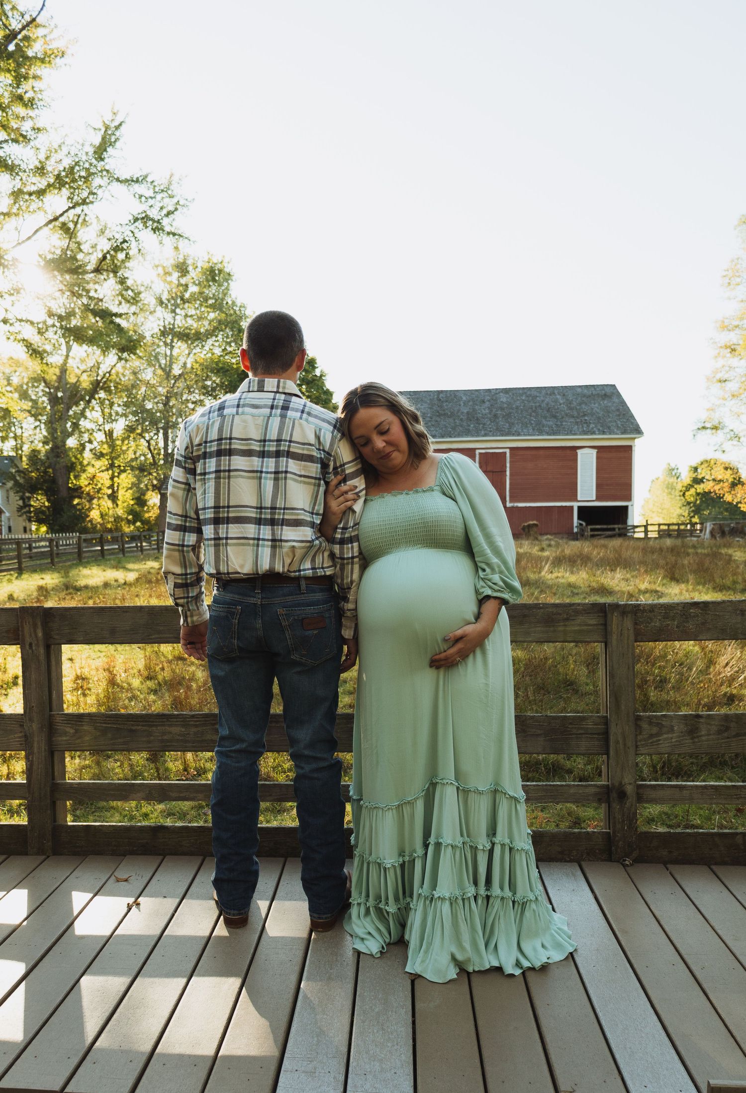 A couple stands together on a wooden deck with a rustic barn and autumn scenery in the background.