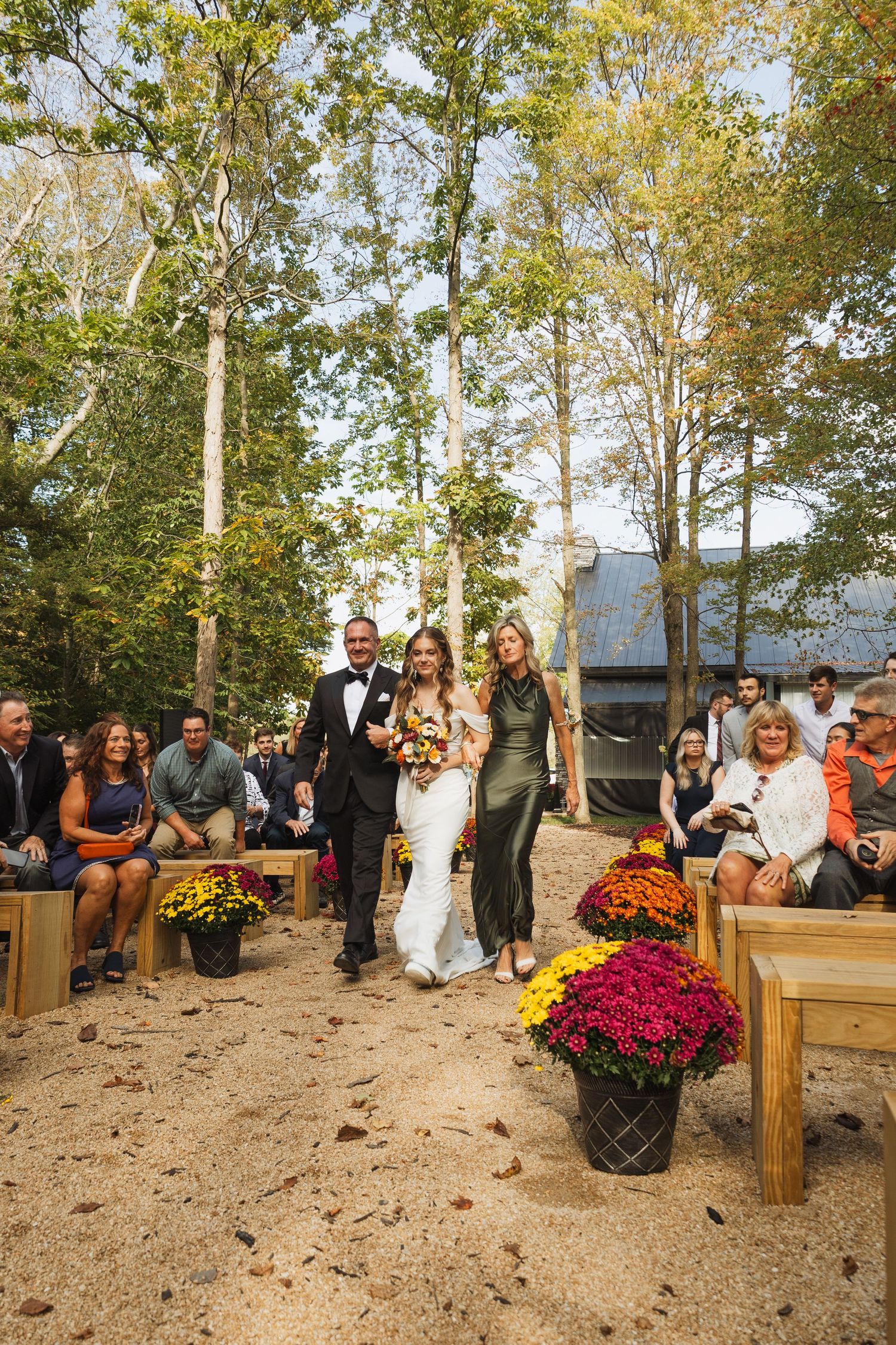 Wedding guests seated on rustic wooden benches surrounded by autumn trees and colorful mums in an outdoor ceremony space.