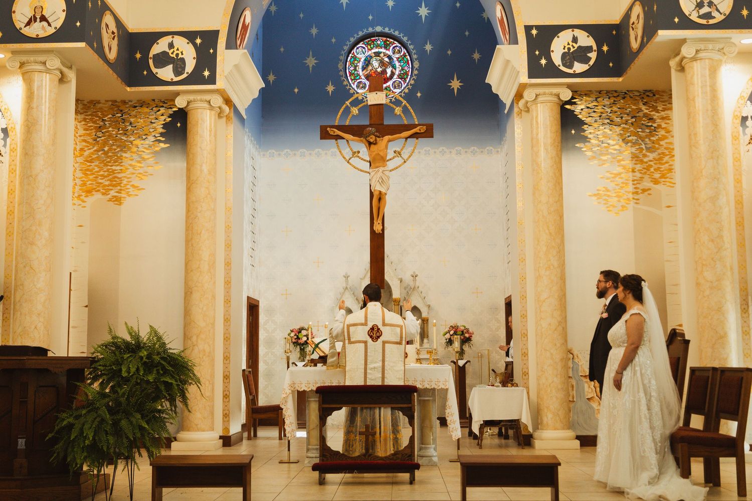 Catholic church altar with ornate crucifix, golden columns and blue starry backdrop during ceremony.