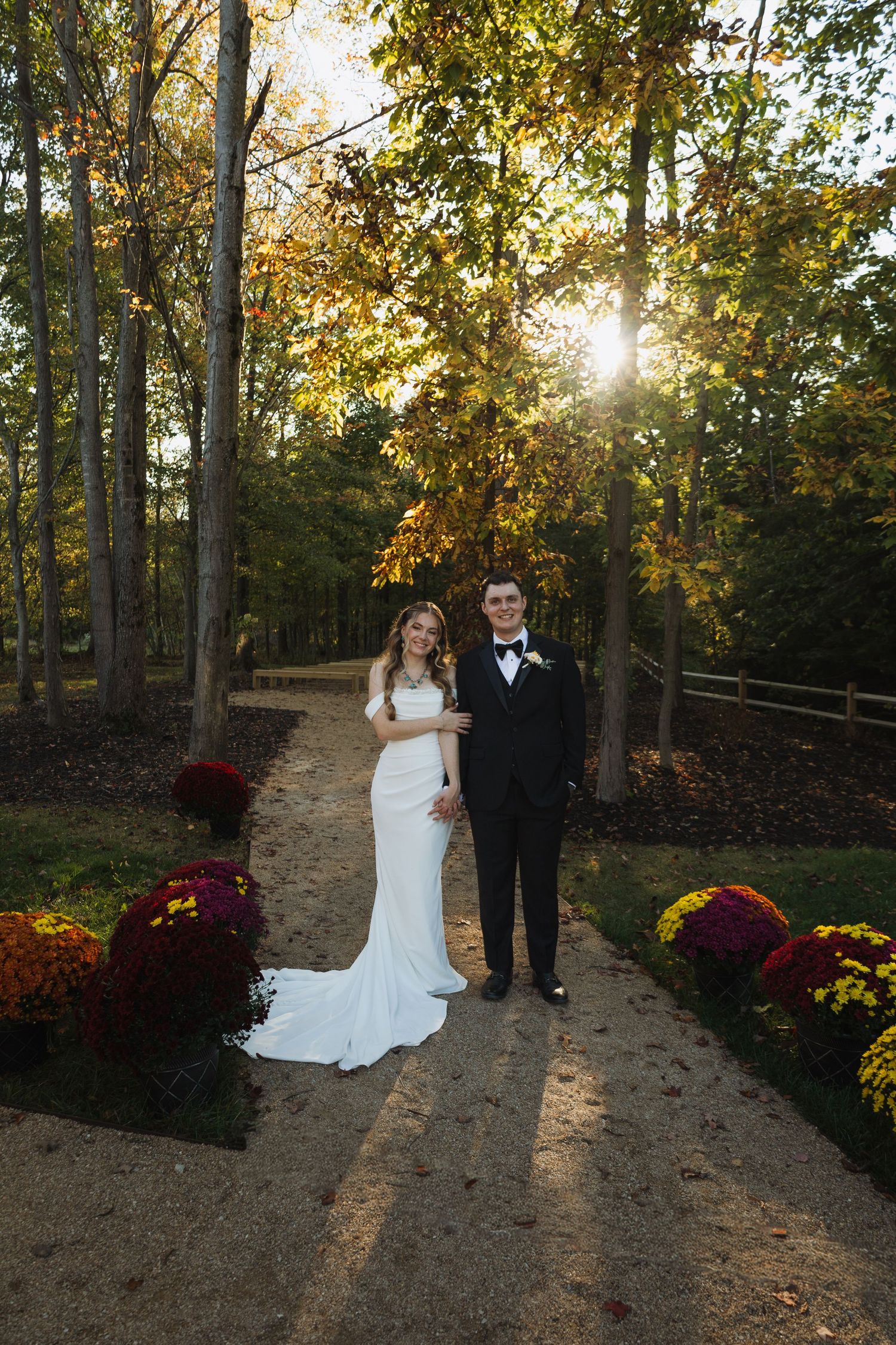 Newlyweds pose on a wooded path with golden sunlight filtering through fall foliage and colorful flowers.