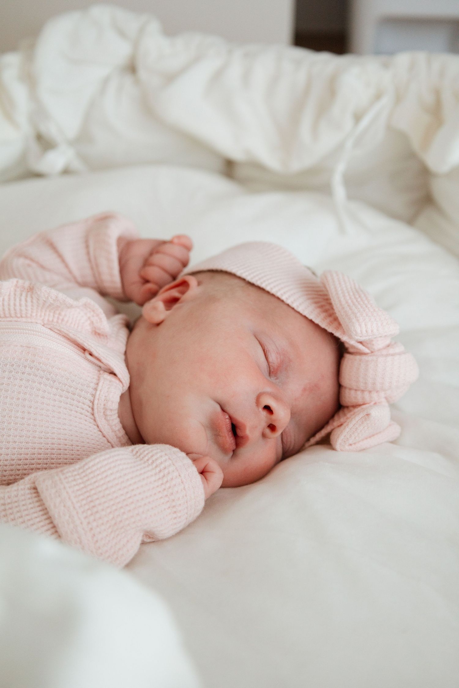 Peaceful sleeping baby wearing soft pink sweater rests on white bedding.