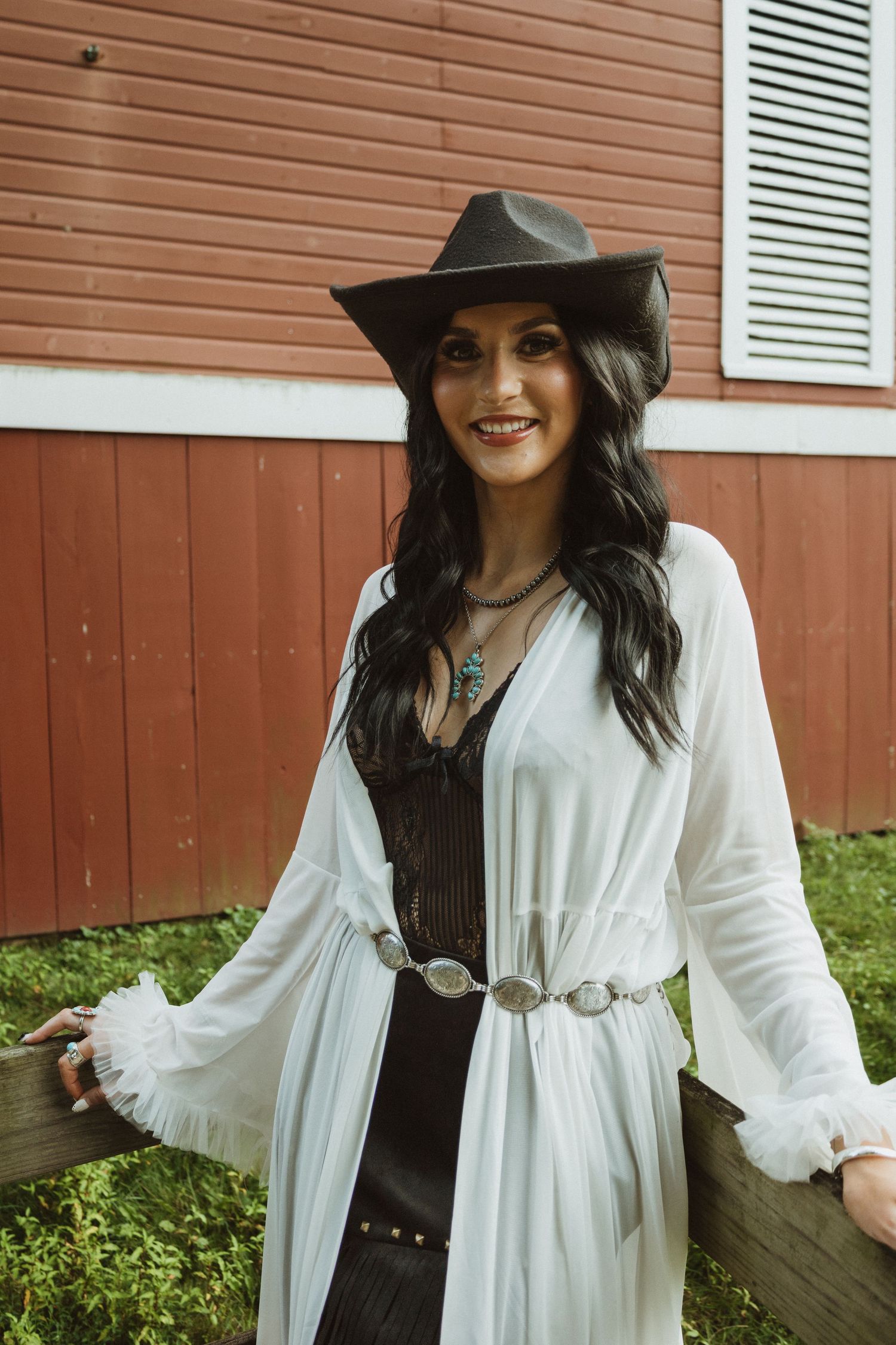 Person wearing a wide-brimmed hat, white flowing dress and western belt poses against a red barn wall.
