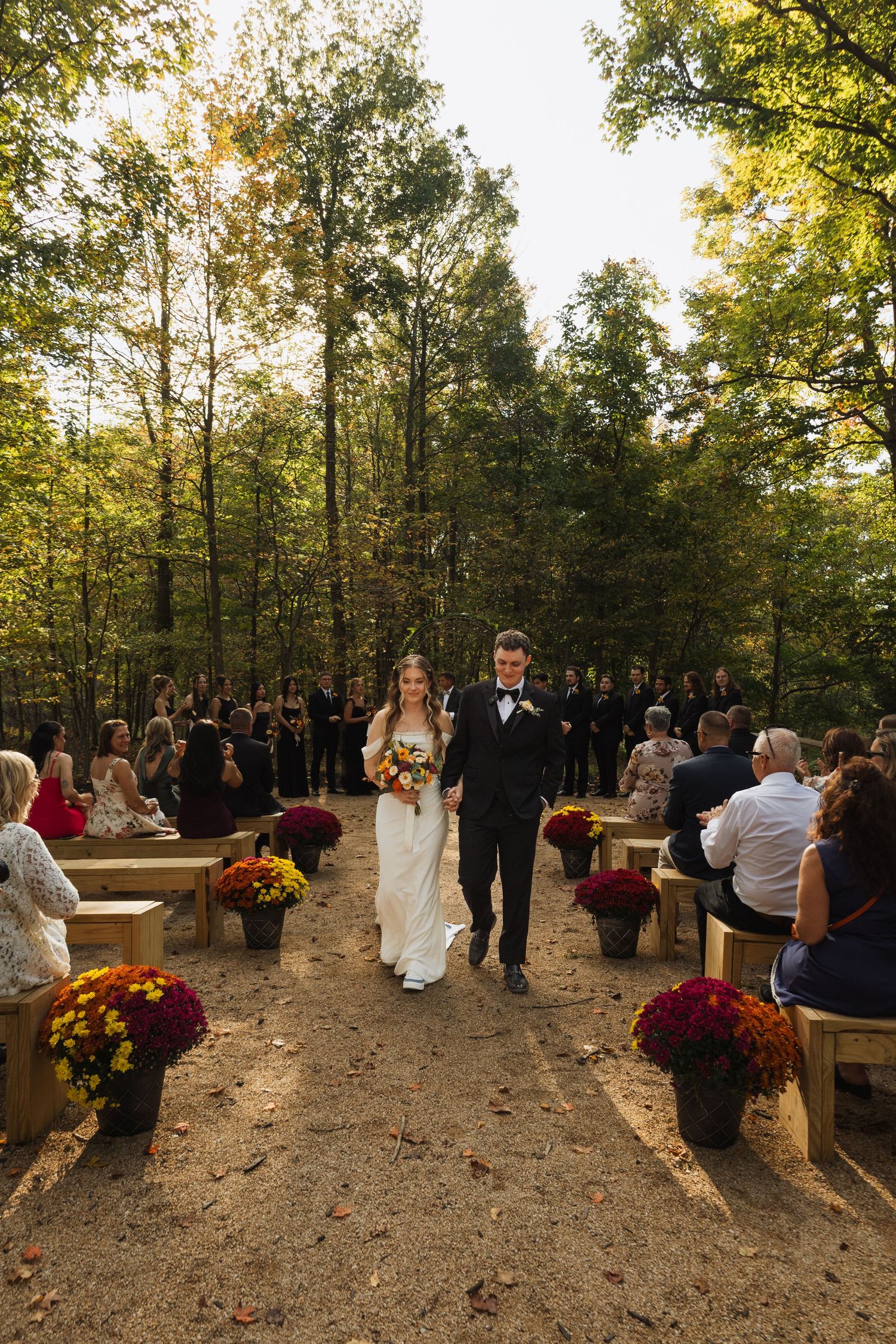Wedding guests gather on rustic benches for an outdoor forest ceremony decorated with fall flowers.