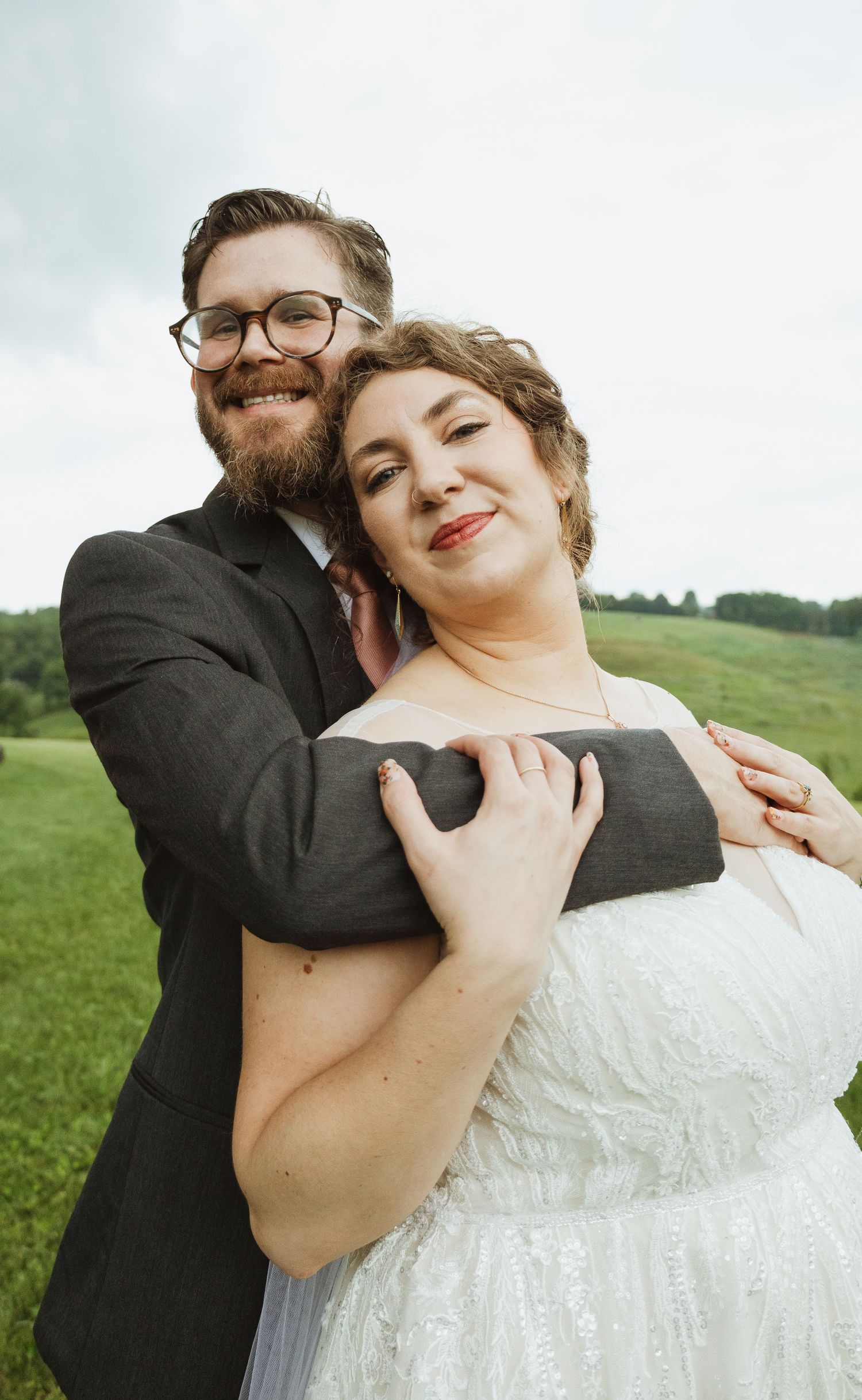 A couple embraces on a green hillside, with the bride wearing a white lace dress and the groom in a dark suit.