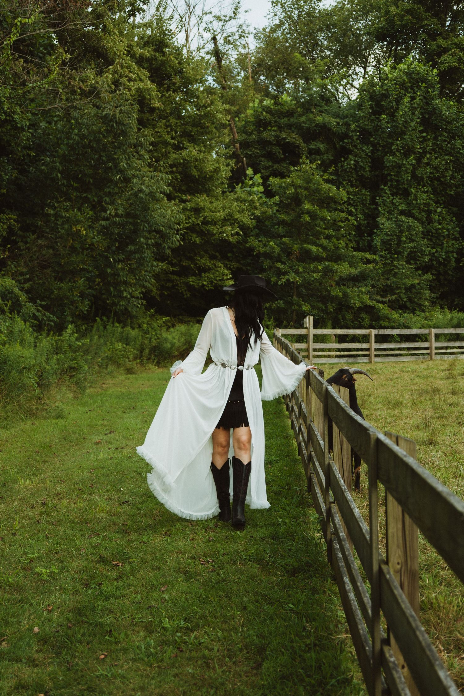 Figure in white dress walks along rustic wooden fence against backdrop of green trees and grass.