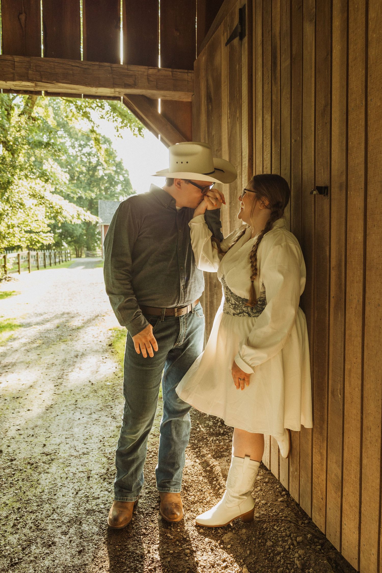 A couple shares a romantic moment against a rustic barn door on a sunlit country path.