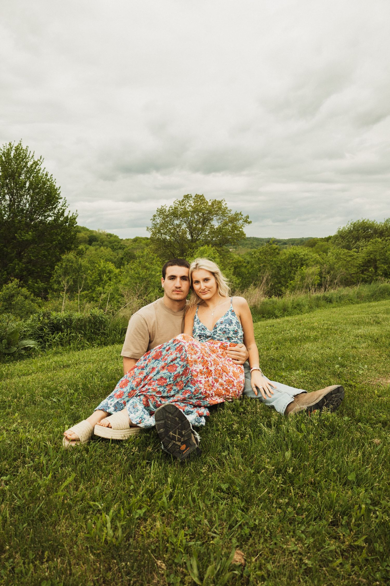 A couple relaxes together on a grassy hillside, with her wearing a floral dress and him in casual attire.