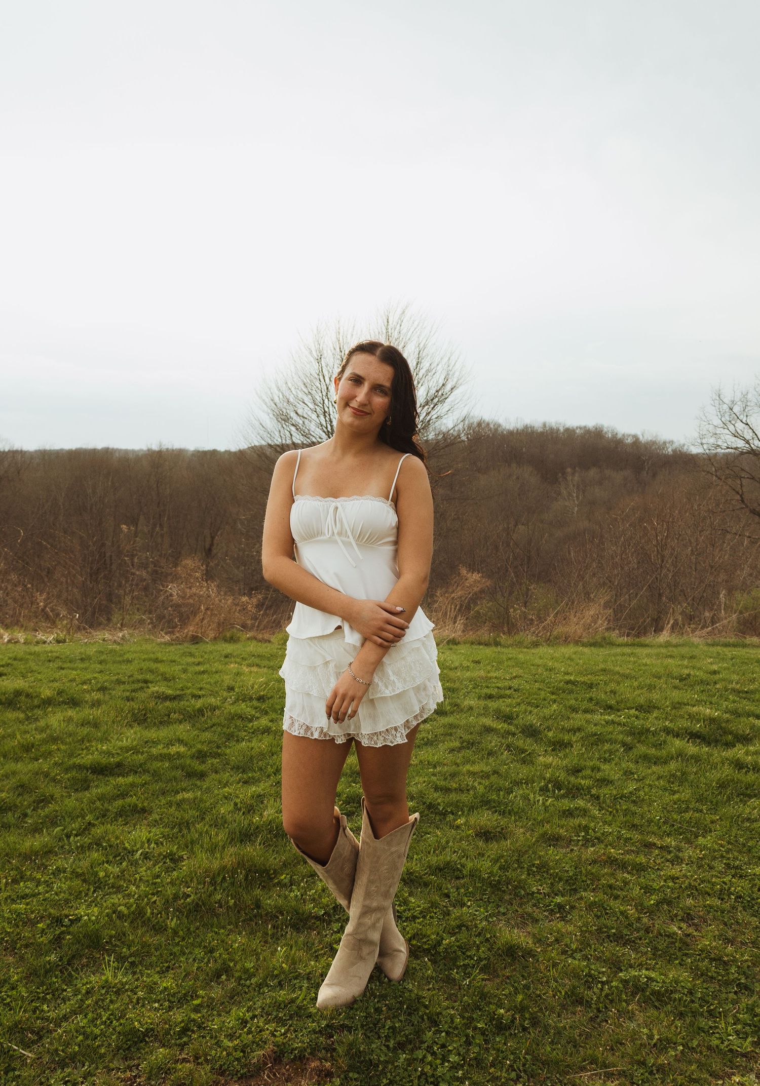 Young woman in white dress and cowboy boots posing in a green field with bare trees in background.