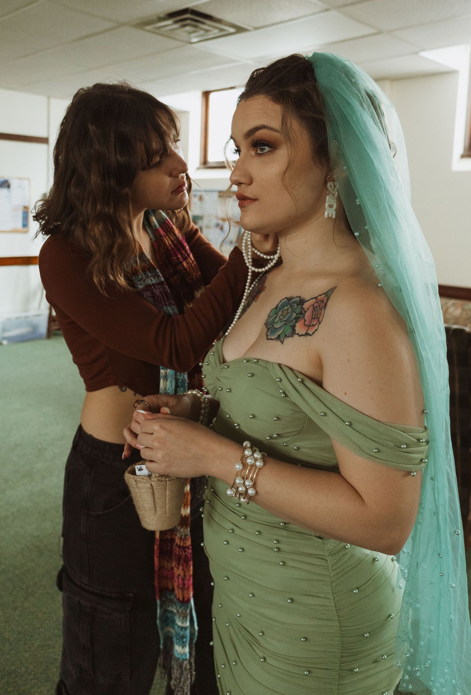 Bridesmaid adjusting green beaded off-shoulder wedding dress on tattooed bride wearing teal veil.