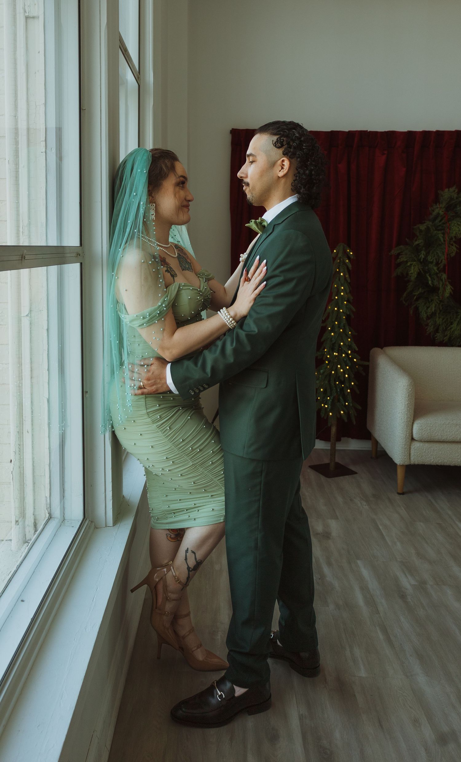Bride and groom in green wedding attire embrace romantically by large windows with Christmas tree in background.