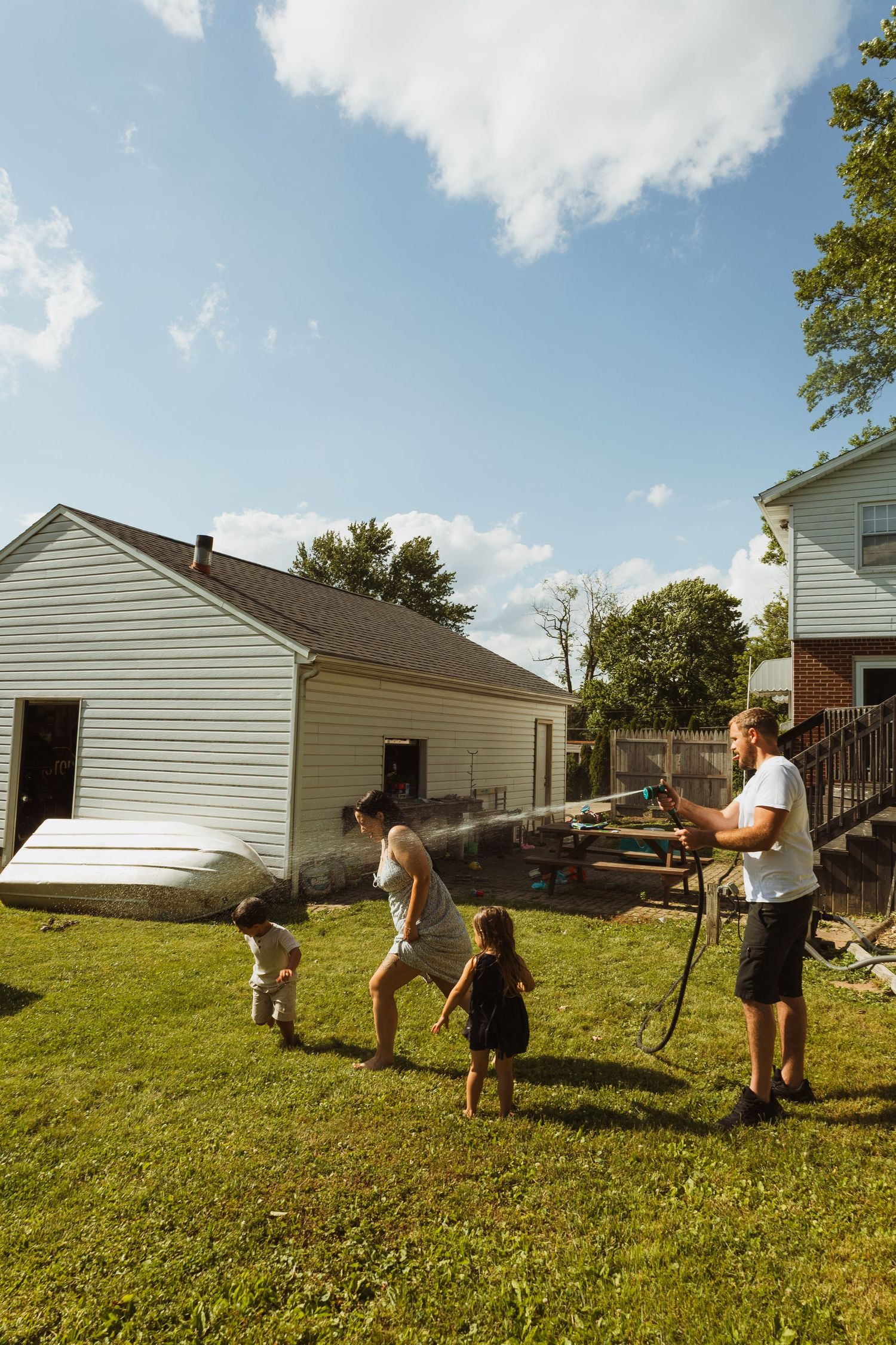 Family playing with garden hose in sunny backyard, parents and two children laughing near white suburban home.