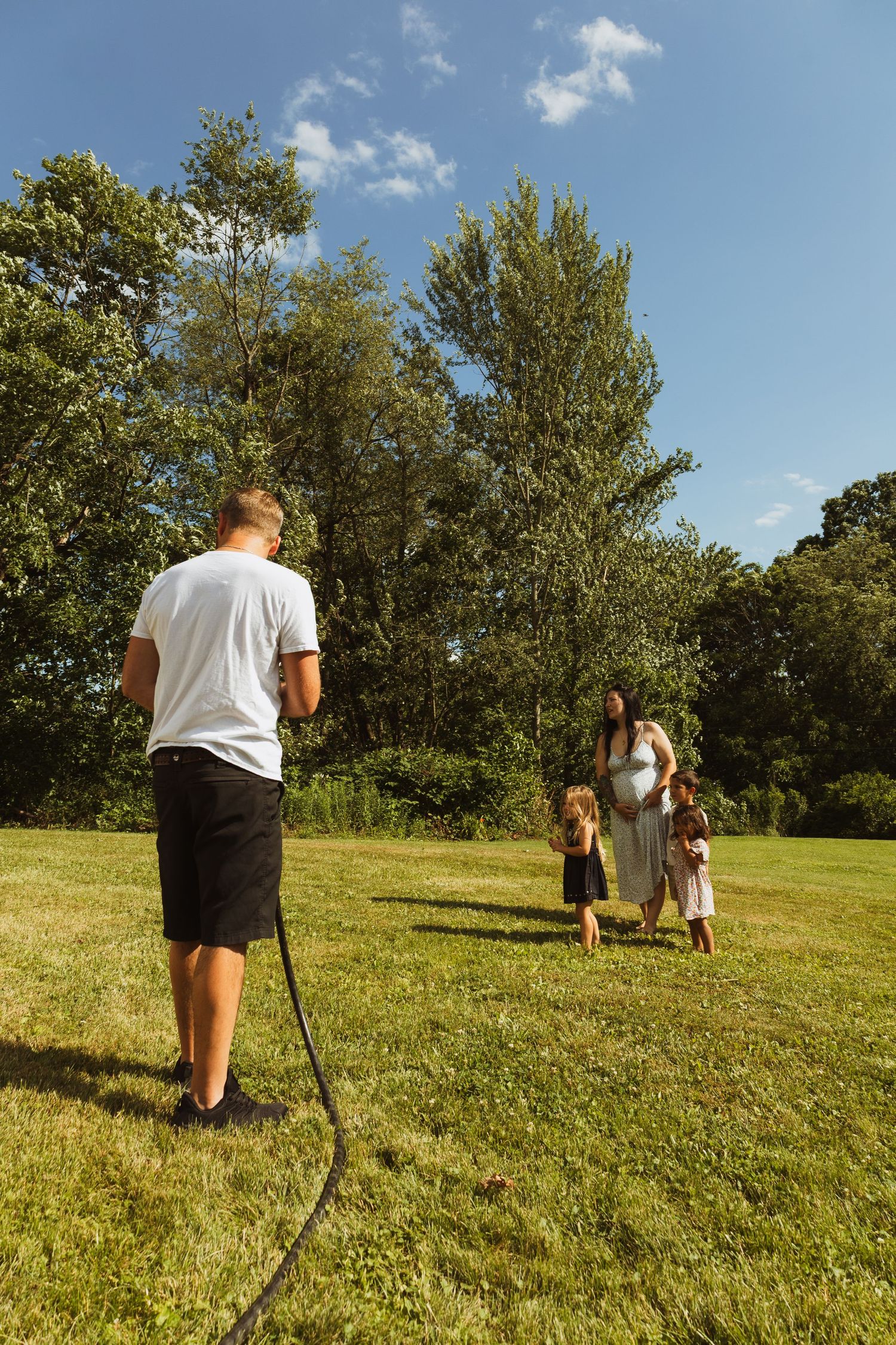 Man with garden hose faces pregnant woman and two children playing on sunny green lawn surrounded by trees.