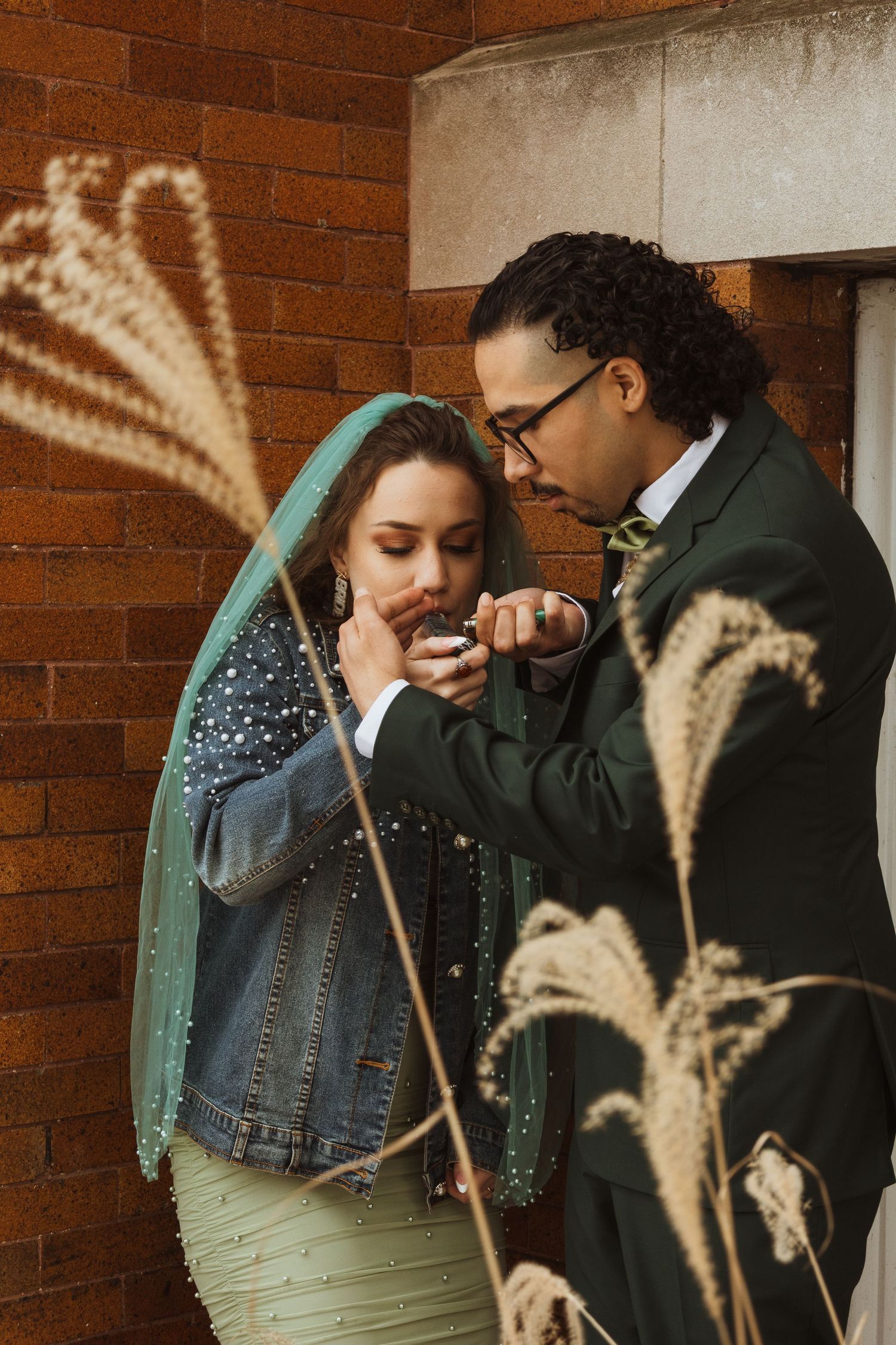Groom in dark green suit tenderly feeds bride in pearl-adorned green dress and veil outdoors.