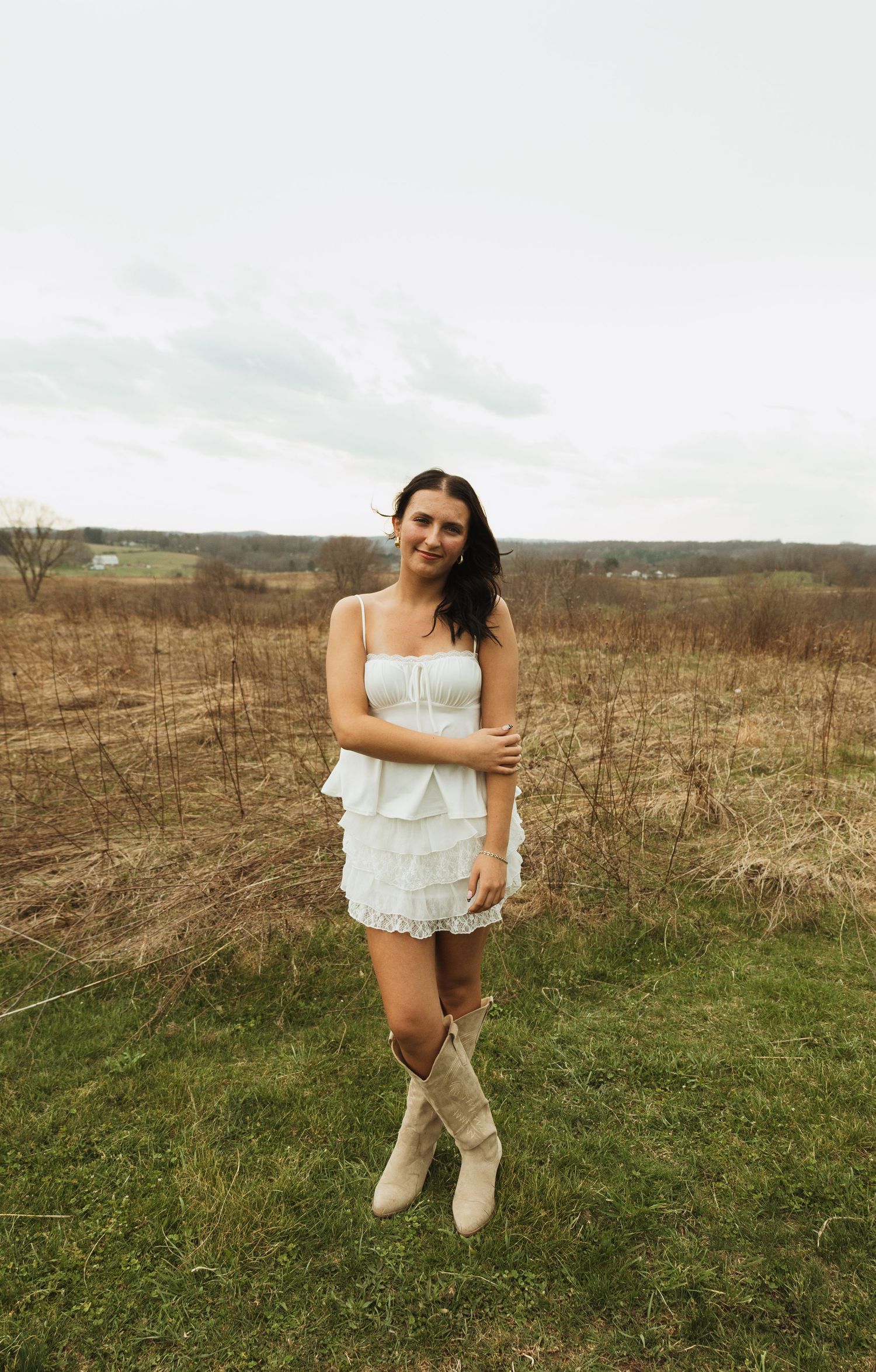 Young woman in white ruffled mini dress and cowboy boots standing in a rustic field under cloudy skies.