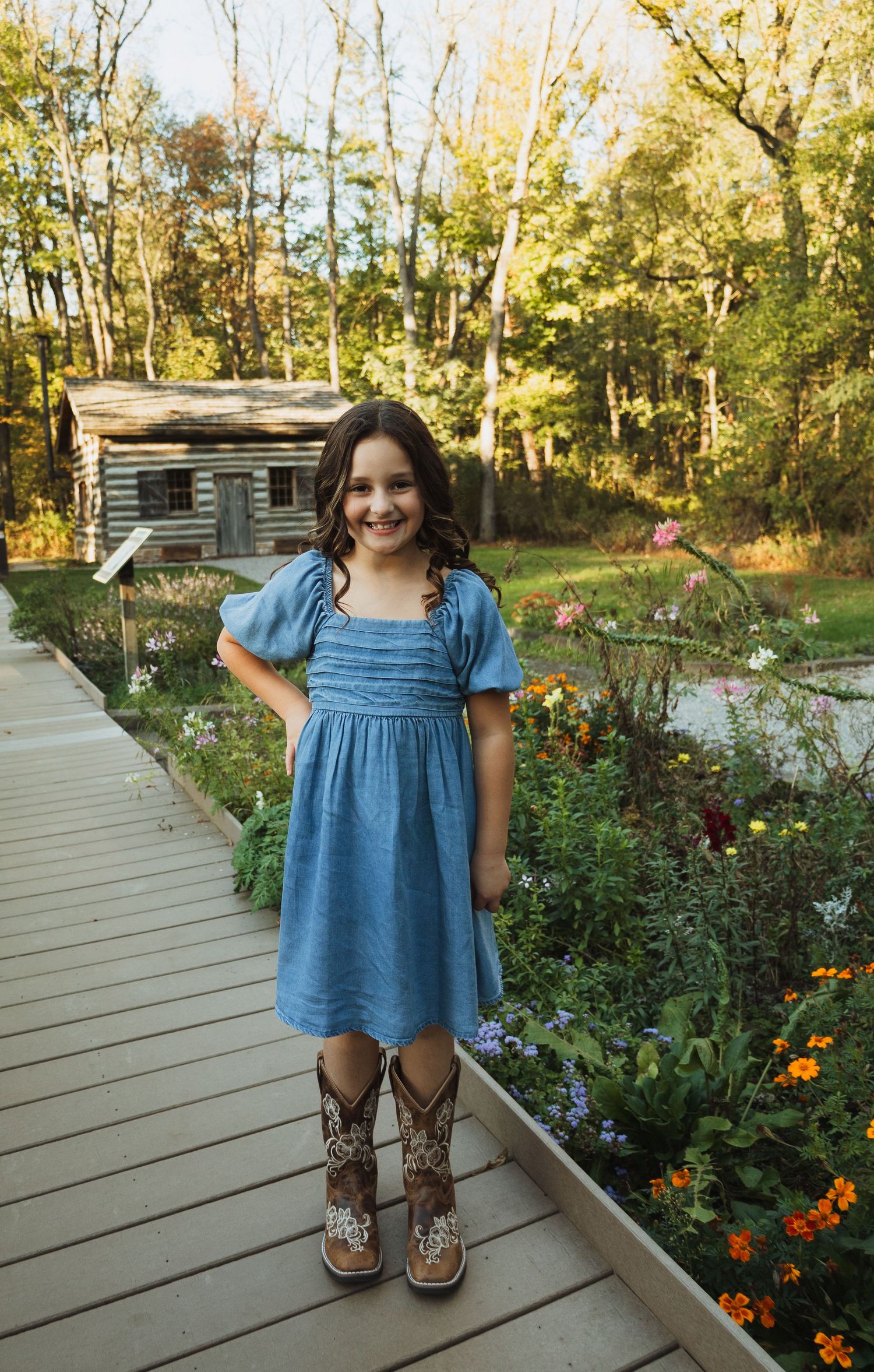 A person in a blue denim dress and cowboy boots stands on a wooden deck surrounded by blooming flowers and trees.