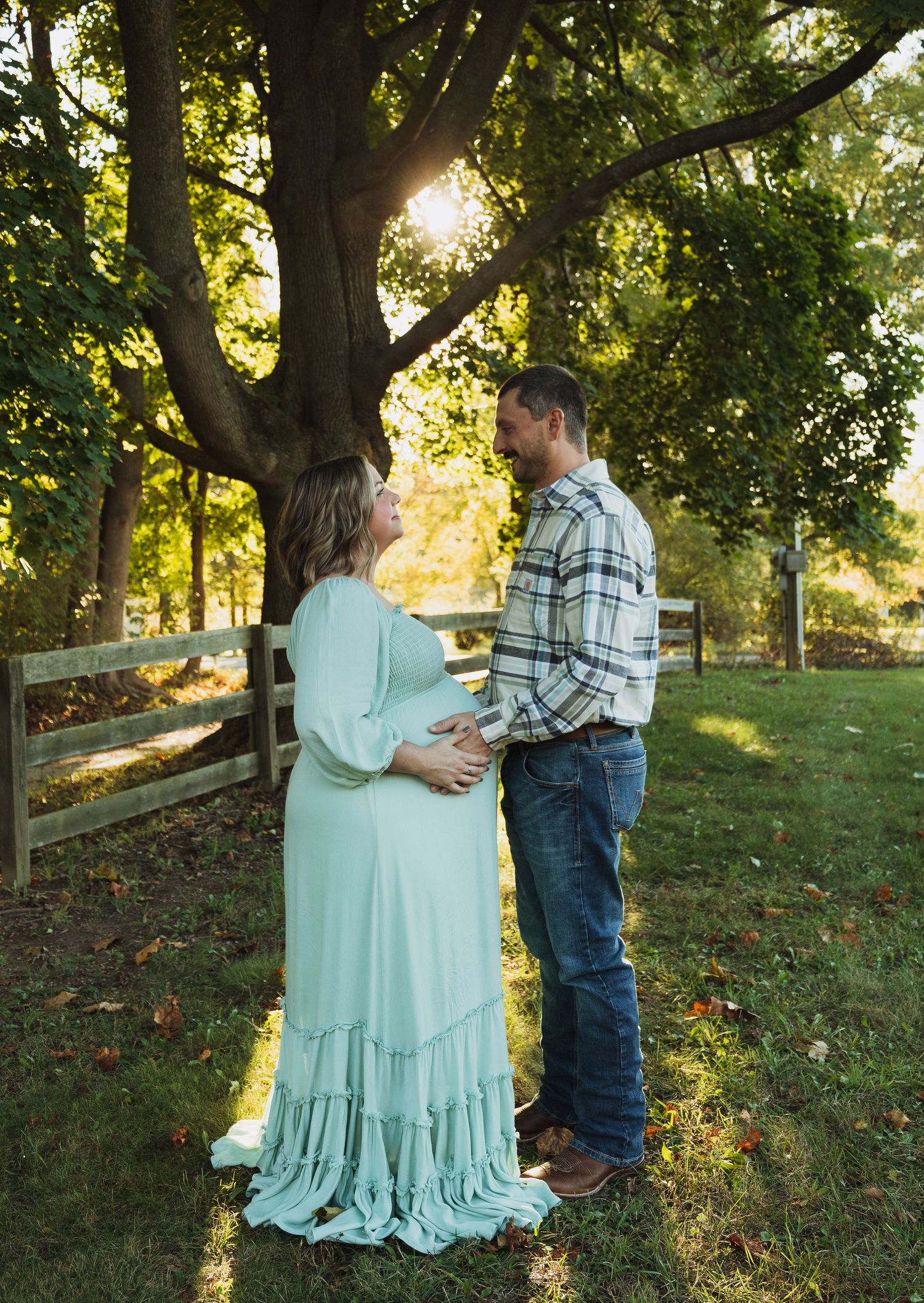 A couple shares a tender moment in a sunlit garden setting with a wooden fence and autumn trees in the background.