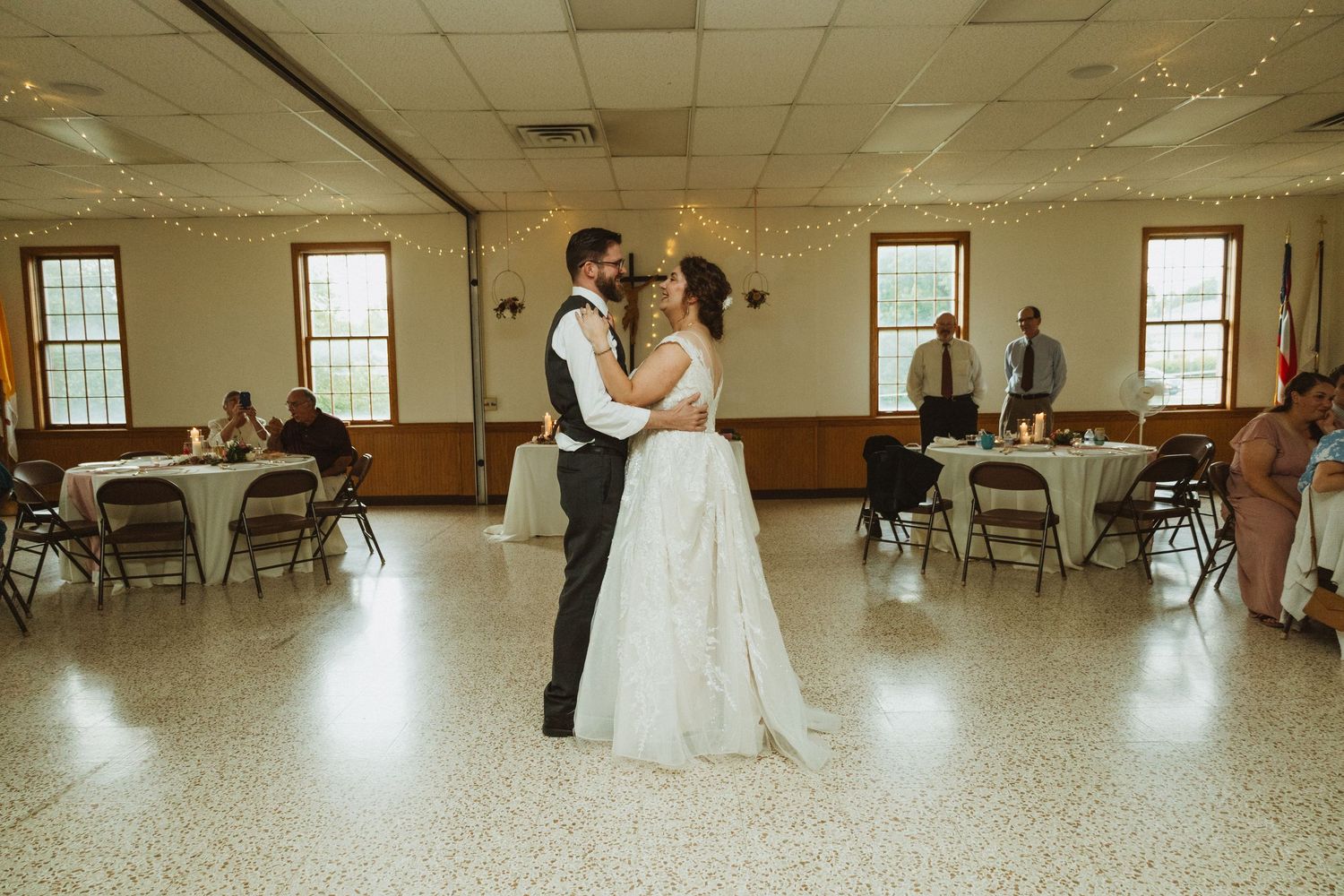 Wedding party dances in a spacious reception hall with white walls and tiled floors.